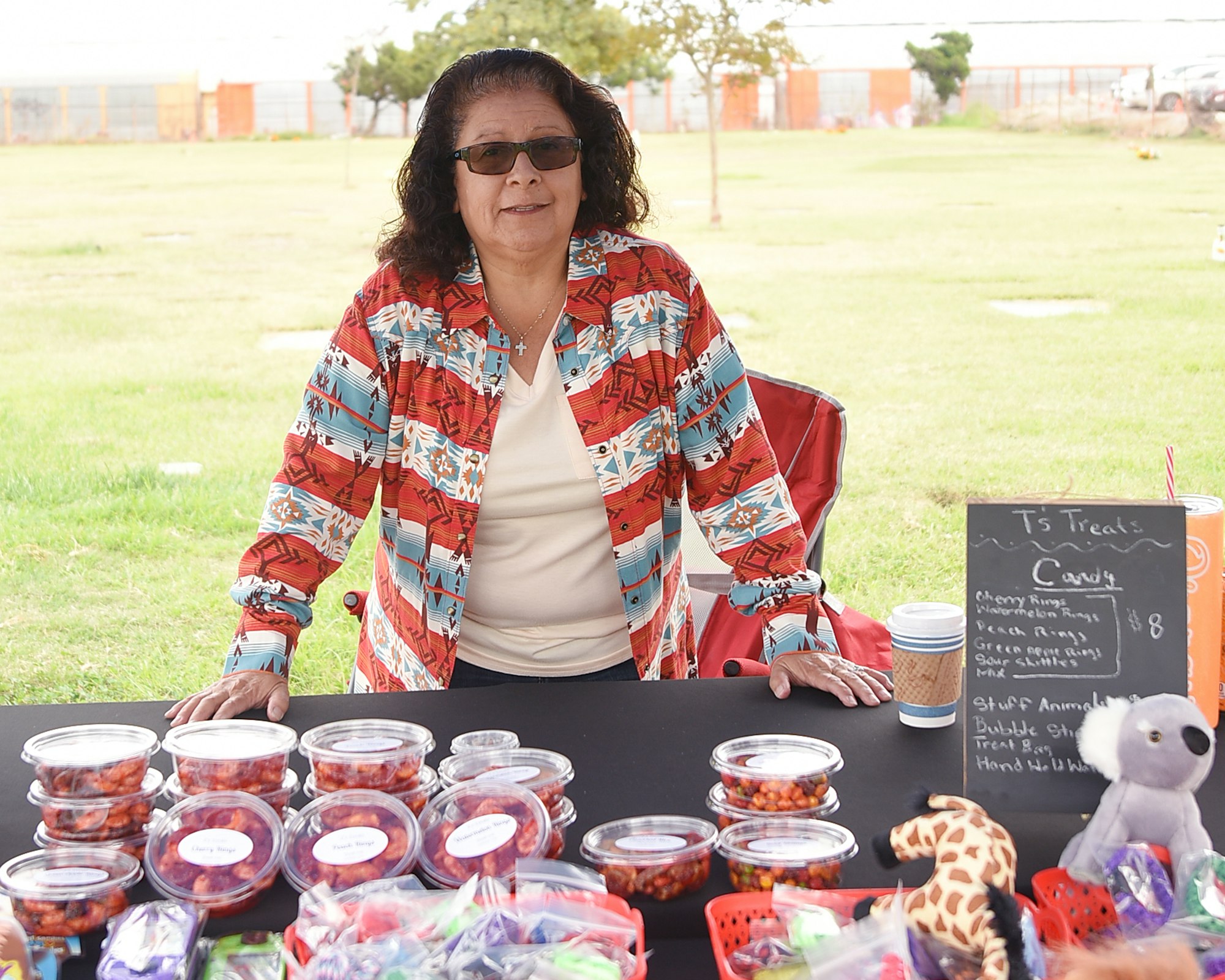 A woman stands behind a table with jars and bags of candy, a sign listing treats, and plush toys, outdoors.