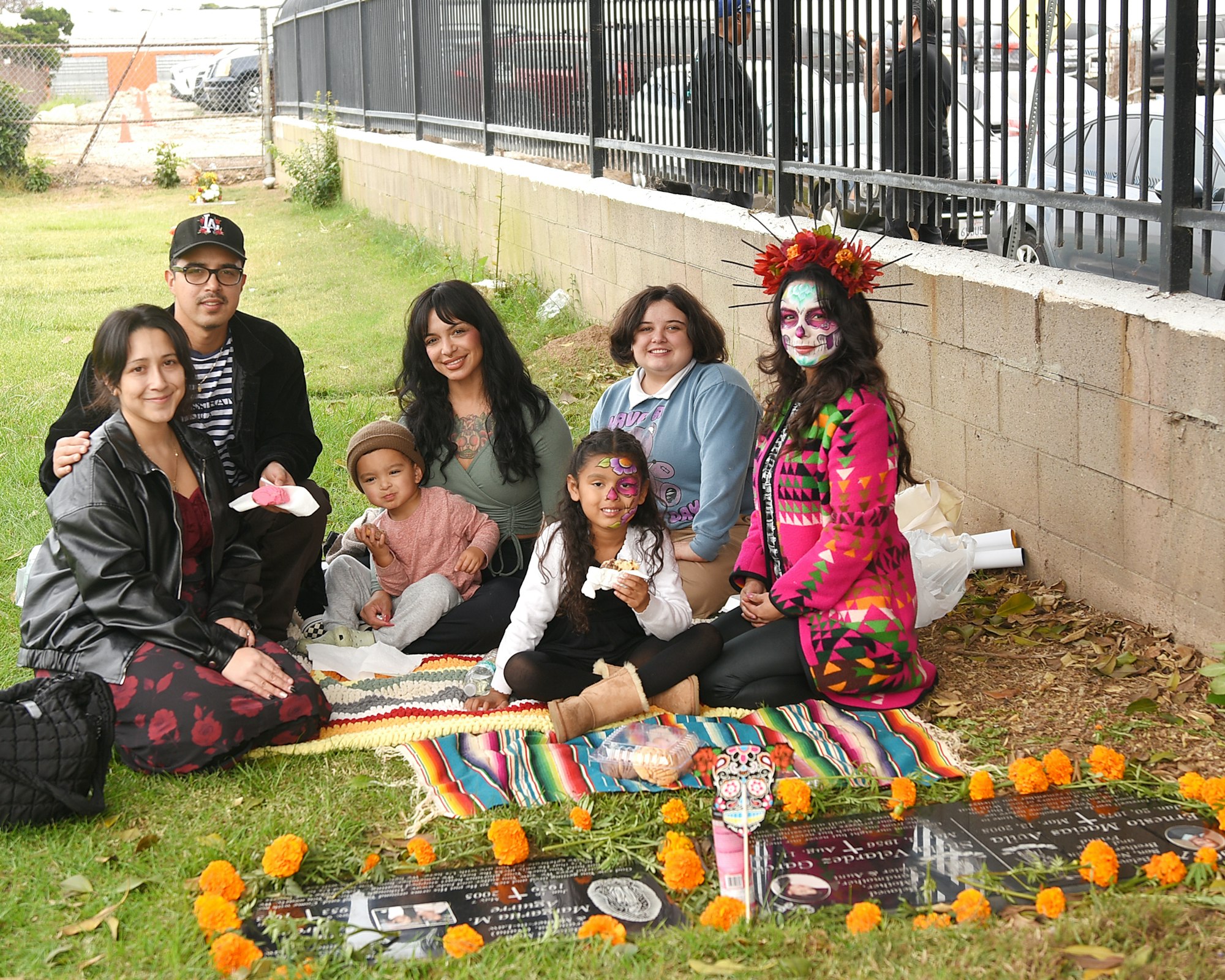 A group of people sitting on the grass with marigolds and a memorial during a cultural celebration. One person wearing Dia de Muertos makeup.