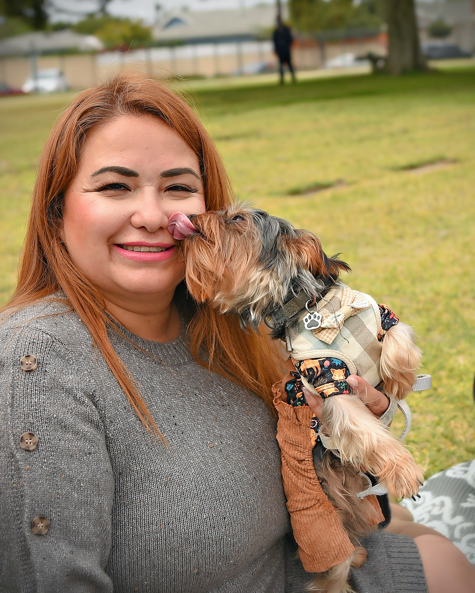 A woman smiling while a small dog licks her face. They're outdoors on a grassy field.