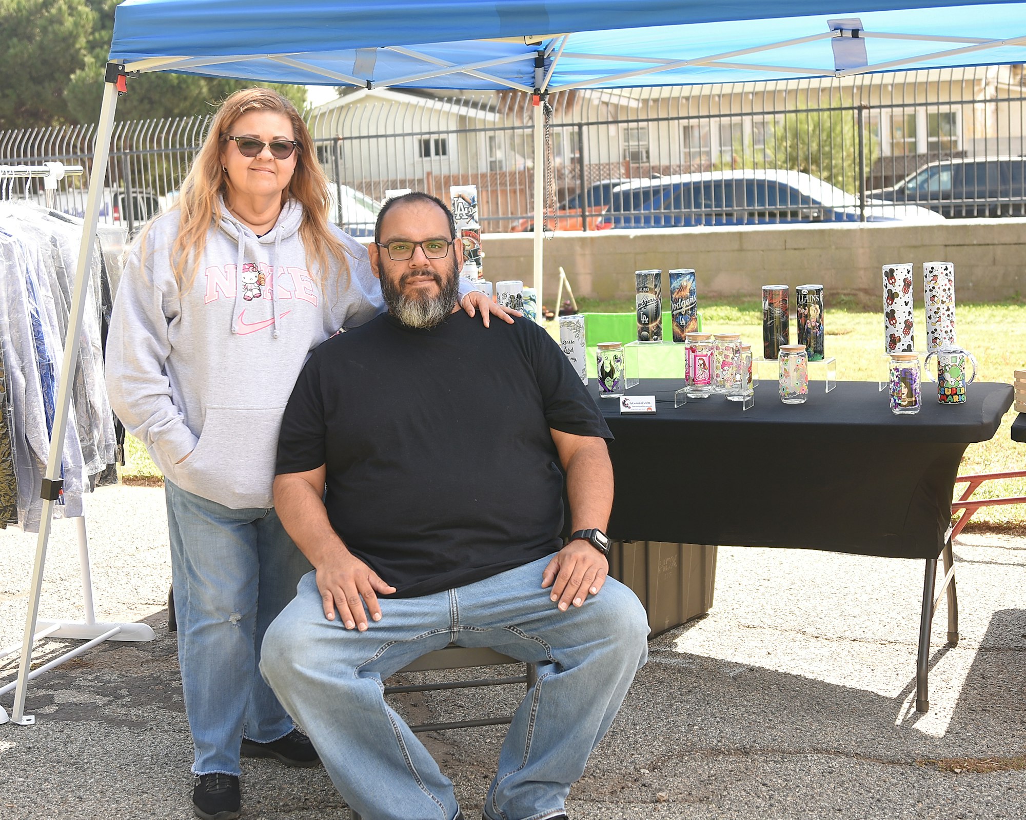 Two people standing by a table with decorative tumblers, under a blue canopy at an outdoor event.