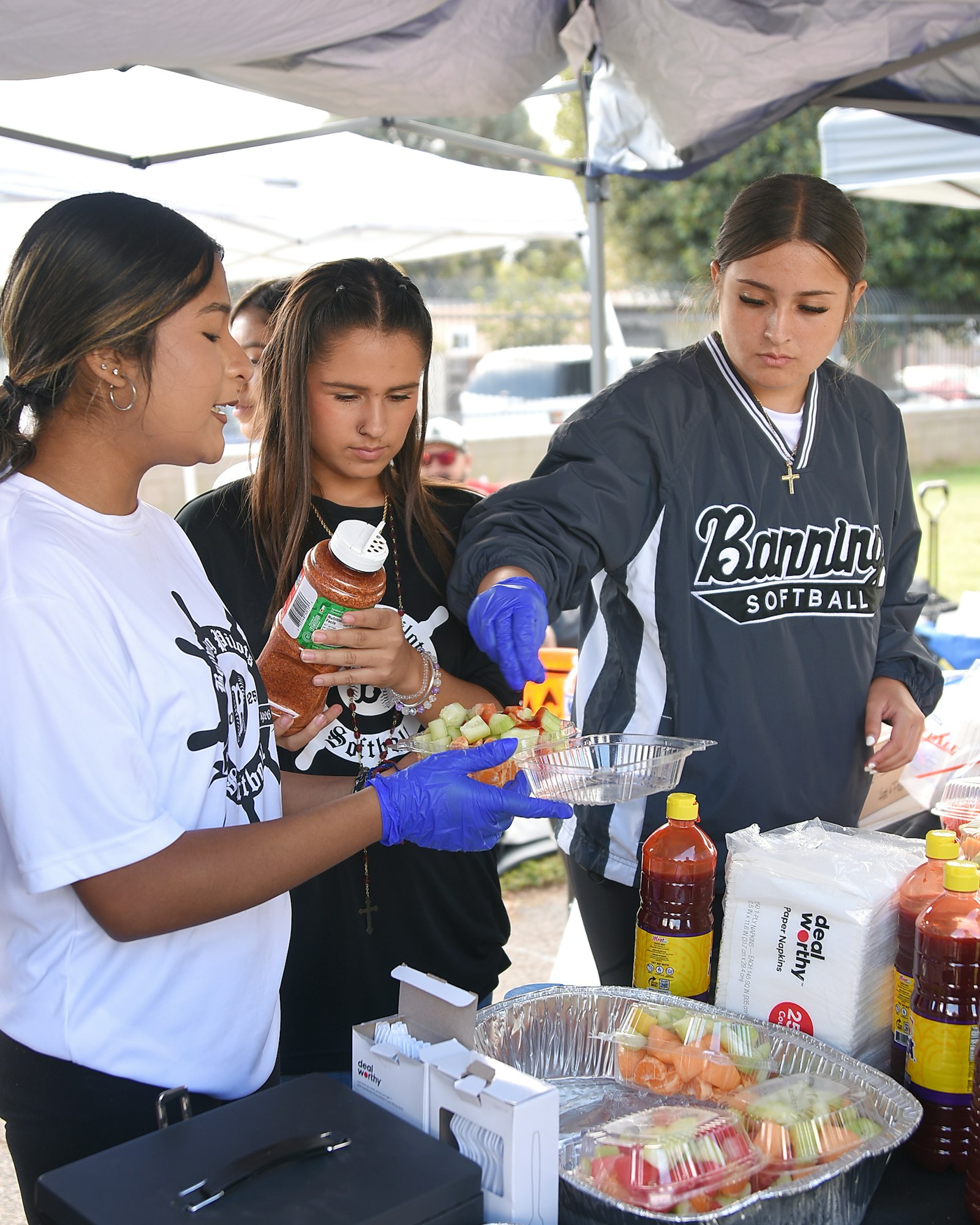 Three people preparing food at a table with fruits and condiments under a tent.
