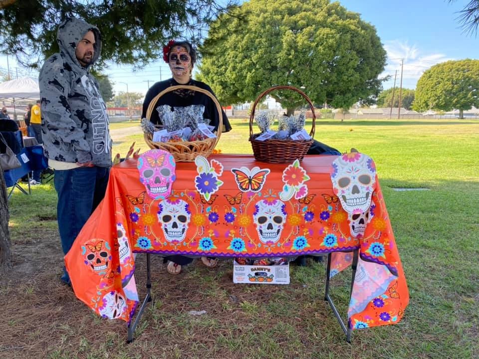 A man and a woman in festive attire are at a decorated table, likely for a cultural event, with a basket and colorful skull designs.