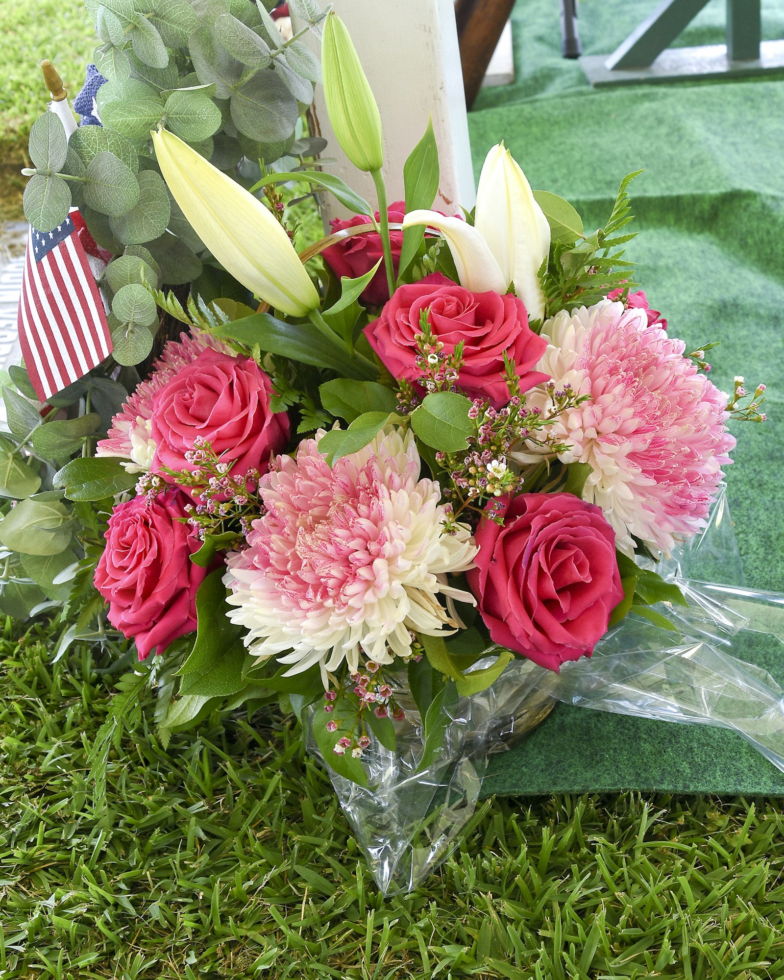 A vibrant bouquet with pink roses, chrysanthemums, lilies, and a small American flag, on a grassy surface.