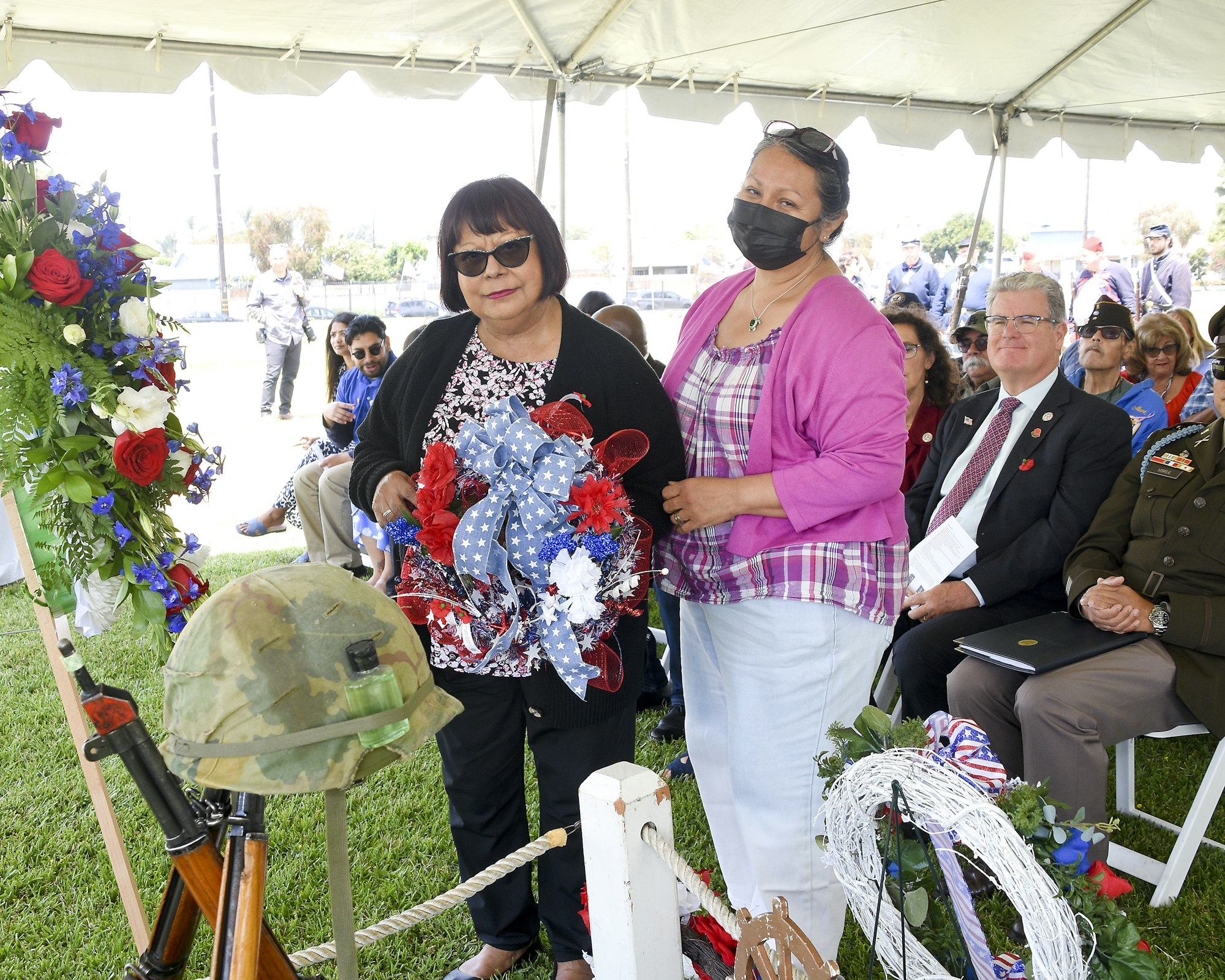 Two women holding a wreath, military helmet on rifle, audience under tent, likely a remembrance or veteran event.