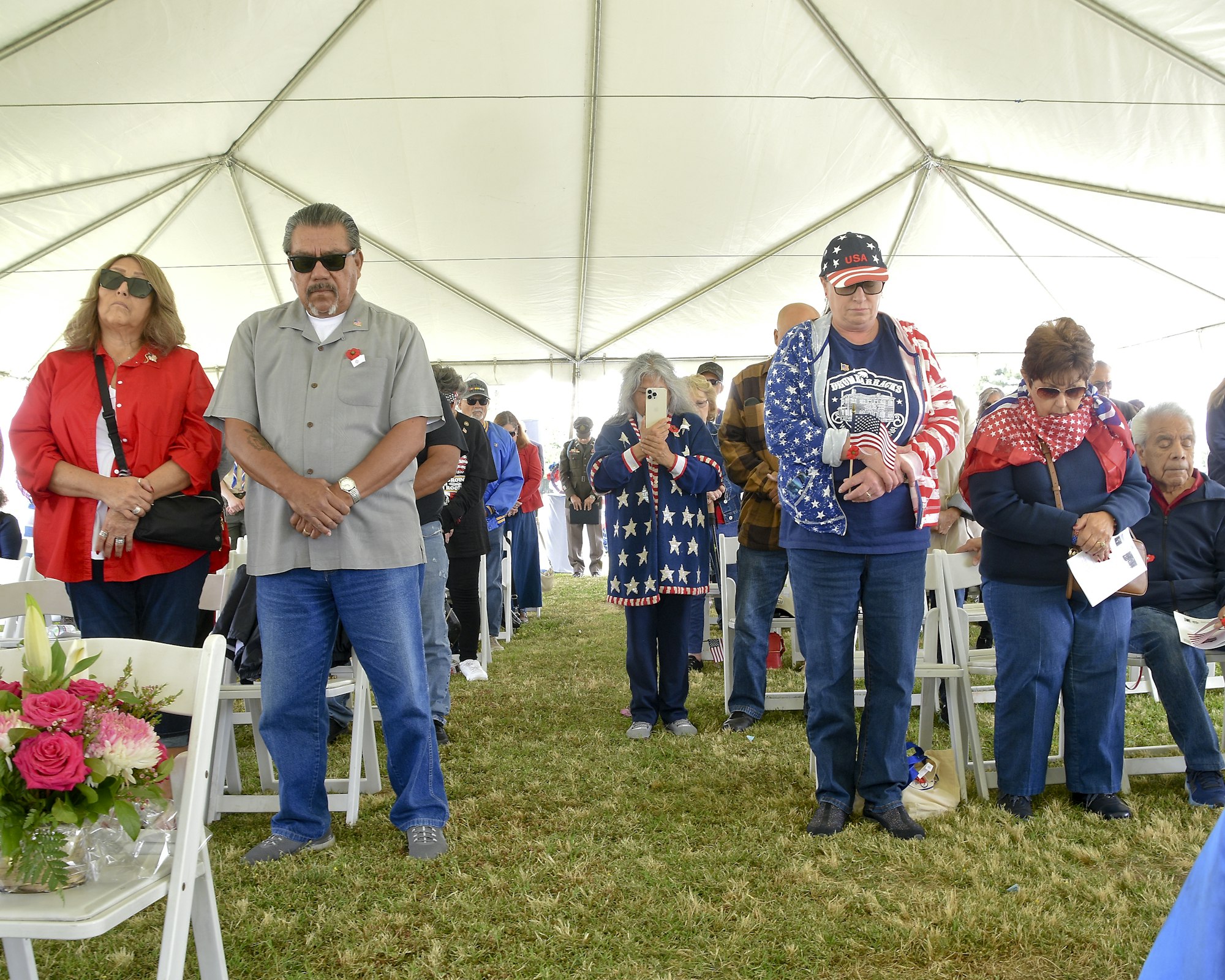 People standing under a tent, possibly at an event, with patriotic clothing and decorations, some with hands over hearts.