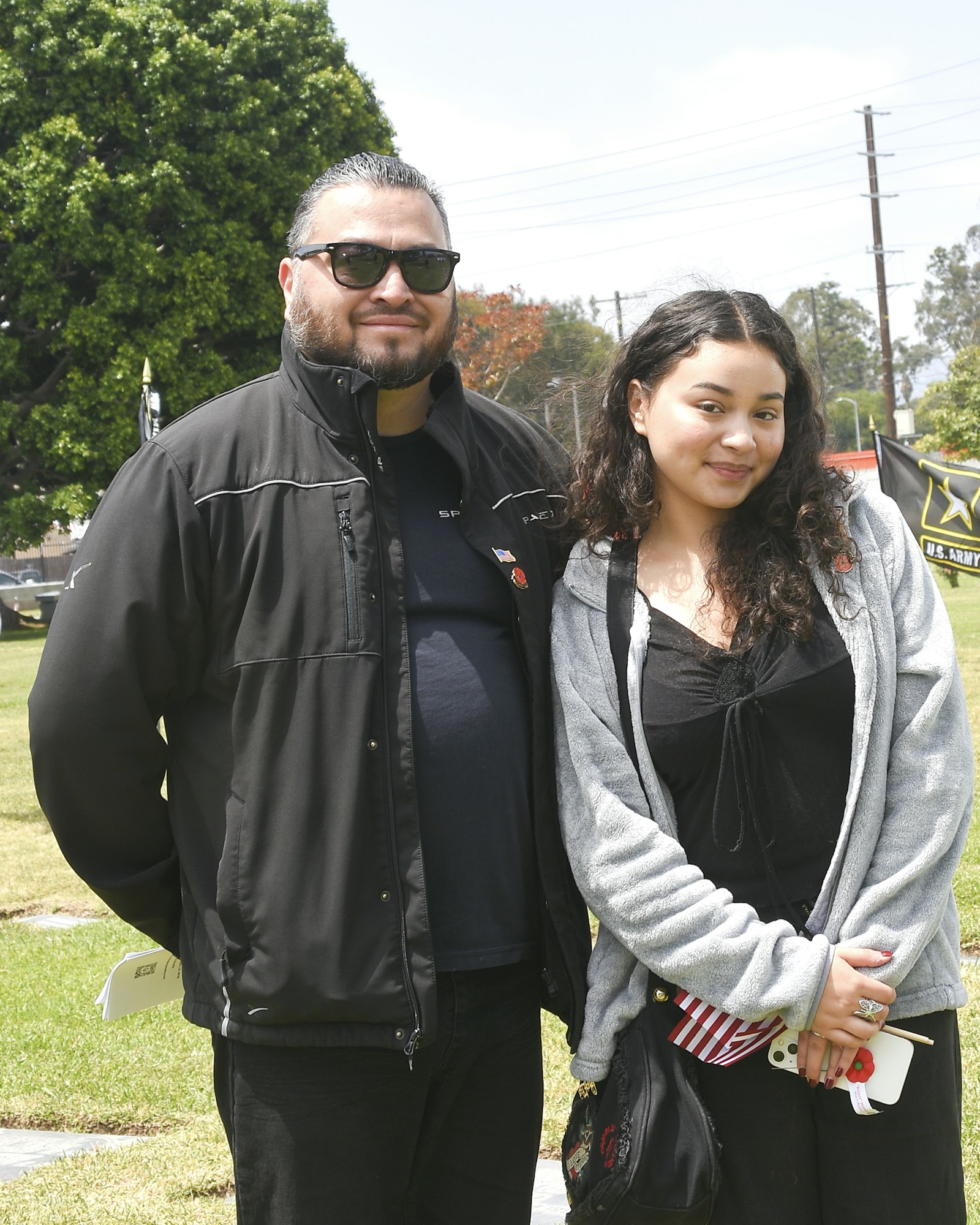 Two people standing outside, likely family, with trees and a flag in the background.