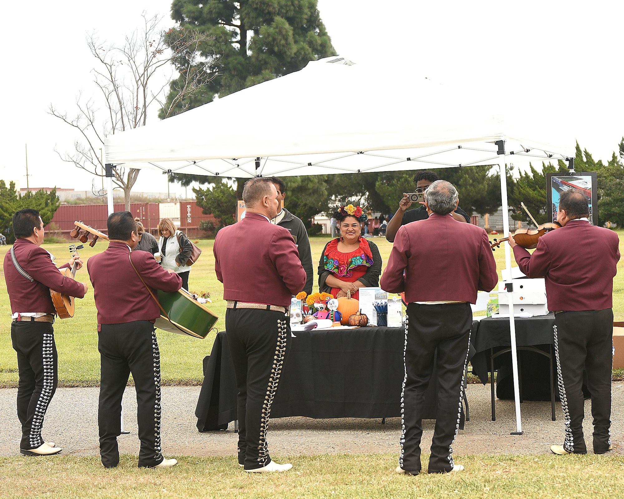 Mariachi band performing outdoors, woman in traditional attire seated, under a canopy, bystanders, greenery, and festive atmosphere.
