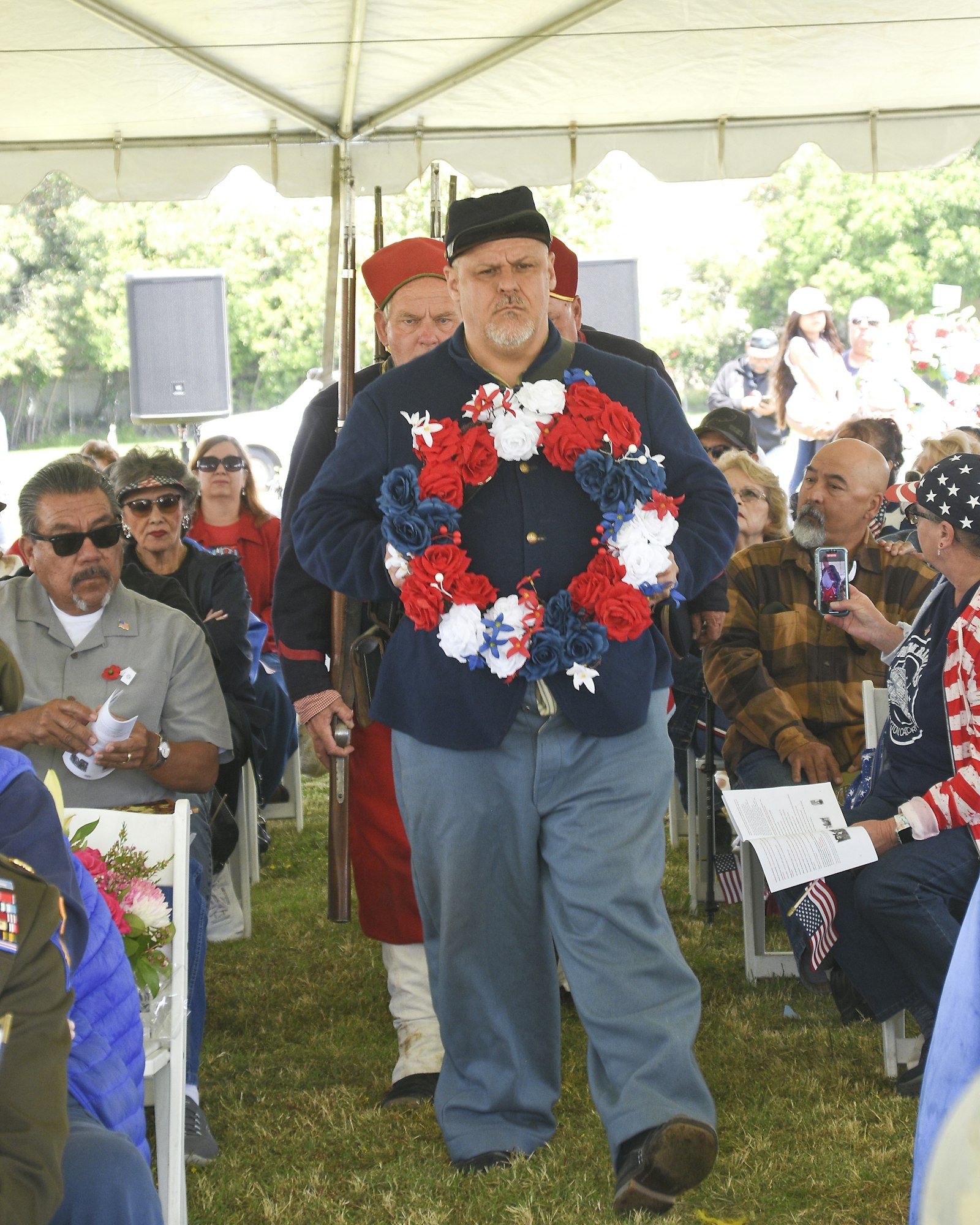 A man holding a wreath at a gathering, possibly a memorial event, with spectators watching.