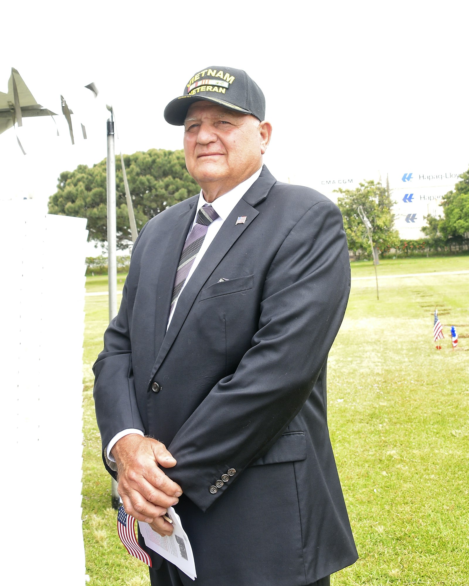 A man wearing a "Vietnam Veteran" cap and suit holds a flag, standing outdoors. There's a monument and trees in the background.