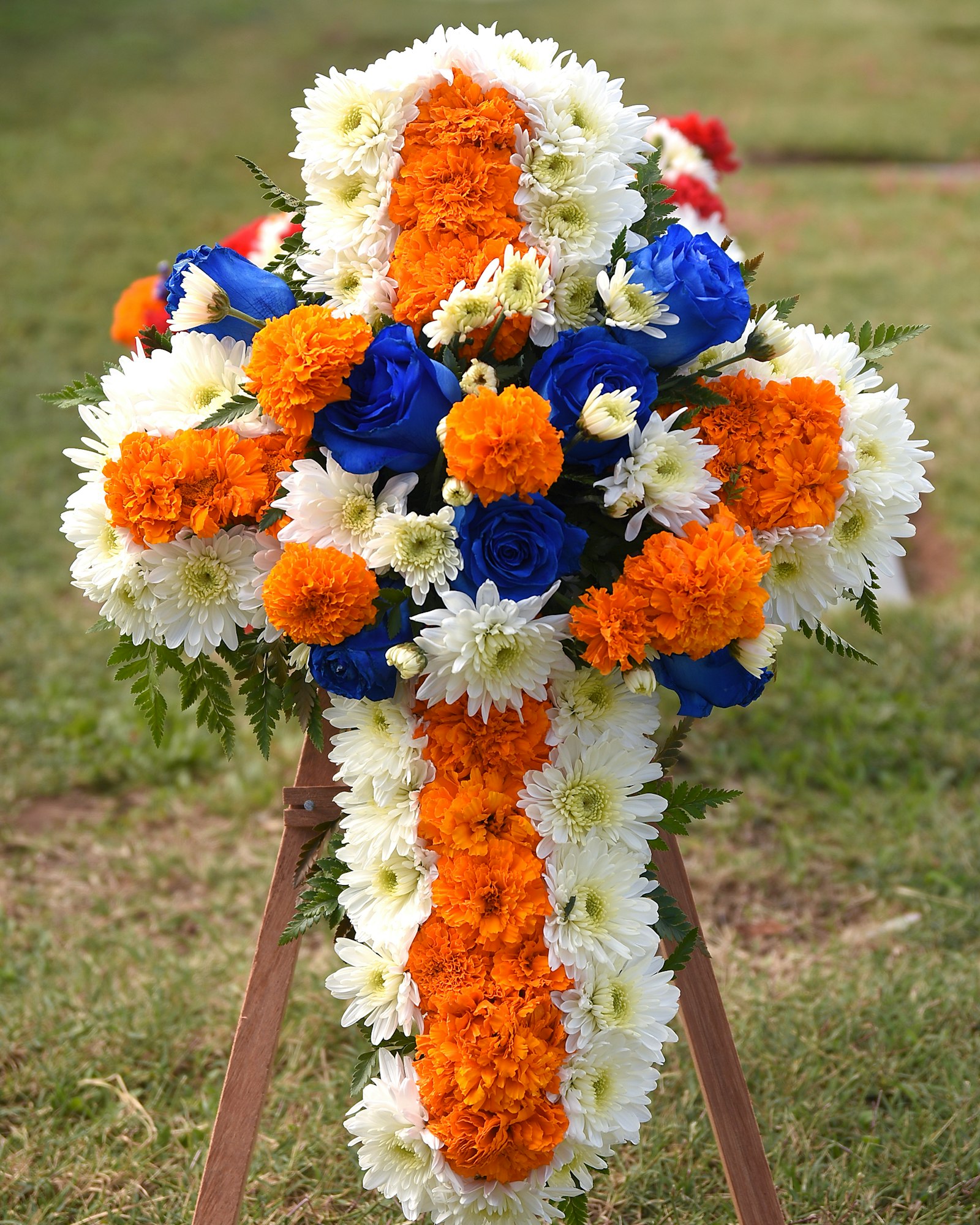A floral display shaped like a cross with orange, white, and blue flowers on a tripod stand.