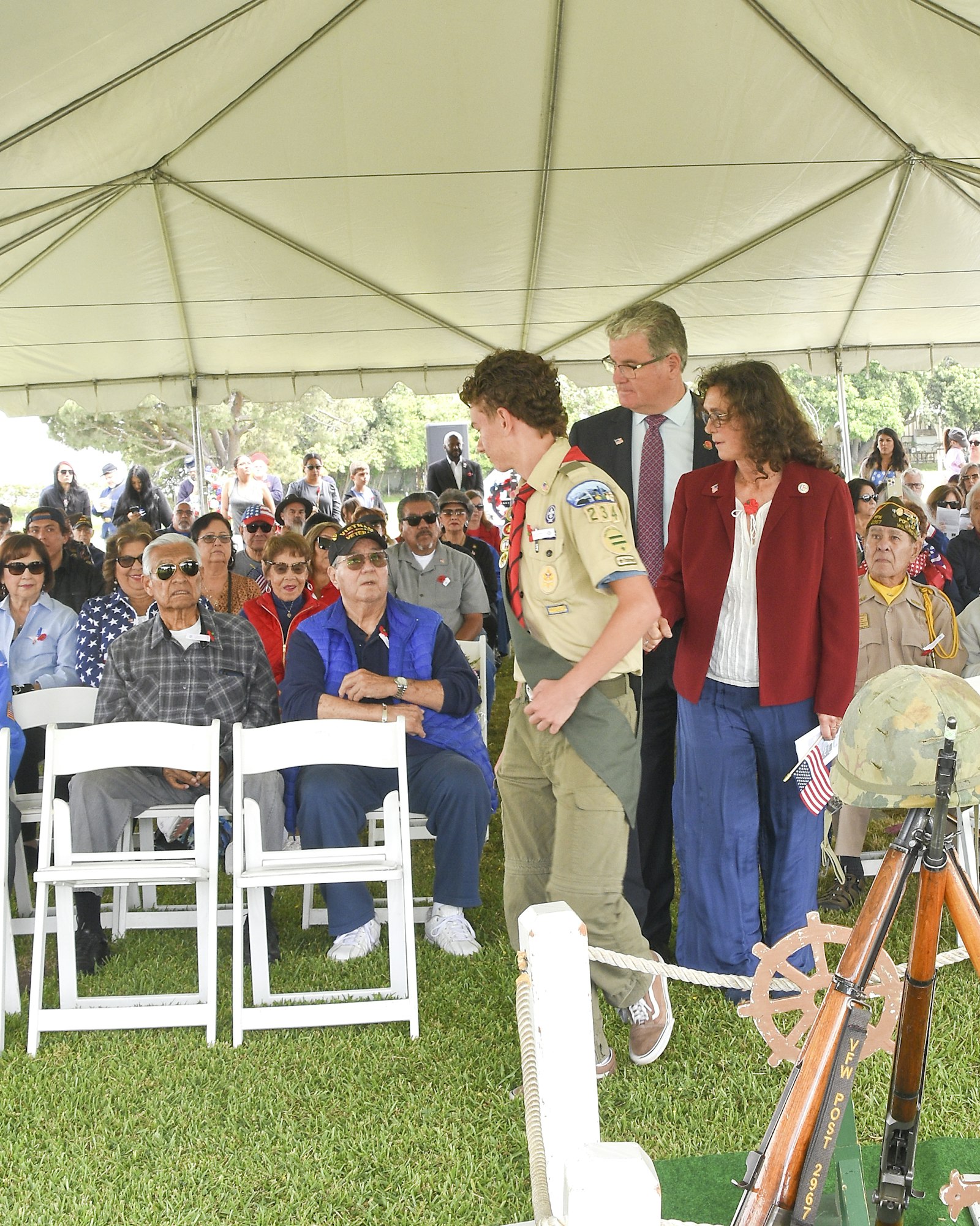 A Boy Scout speaking under a tent while a crowd watches; two adults stand beside him, possibly at a ceremony or event.