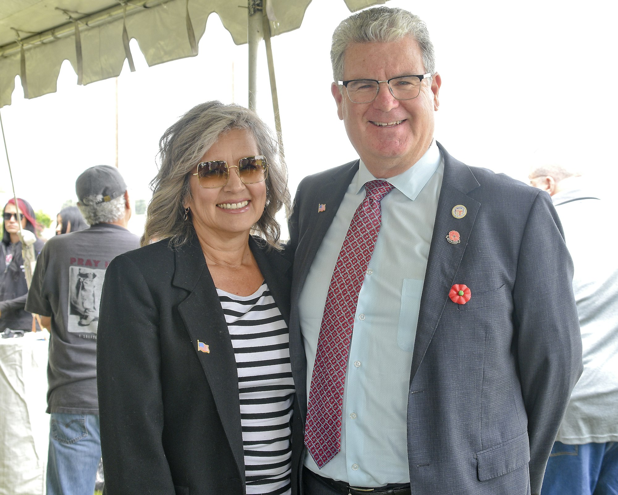 A smiling man and woman posing together, both wearing business attire with patriotic pins. Outdoor setting with tent and people in the background.