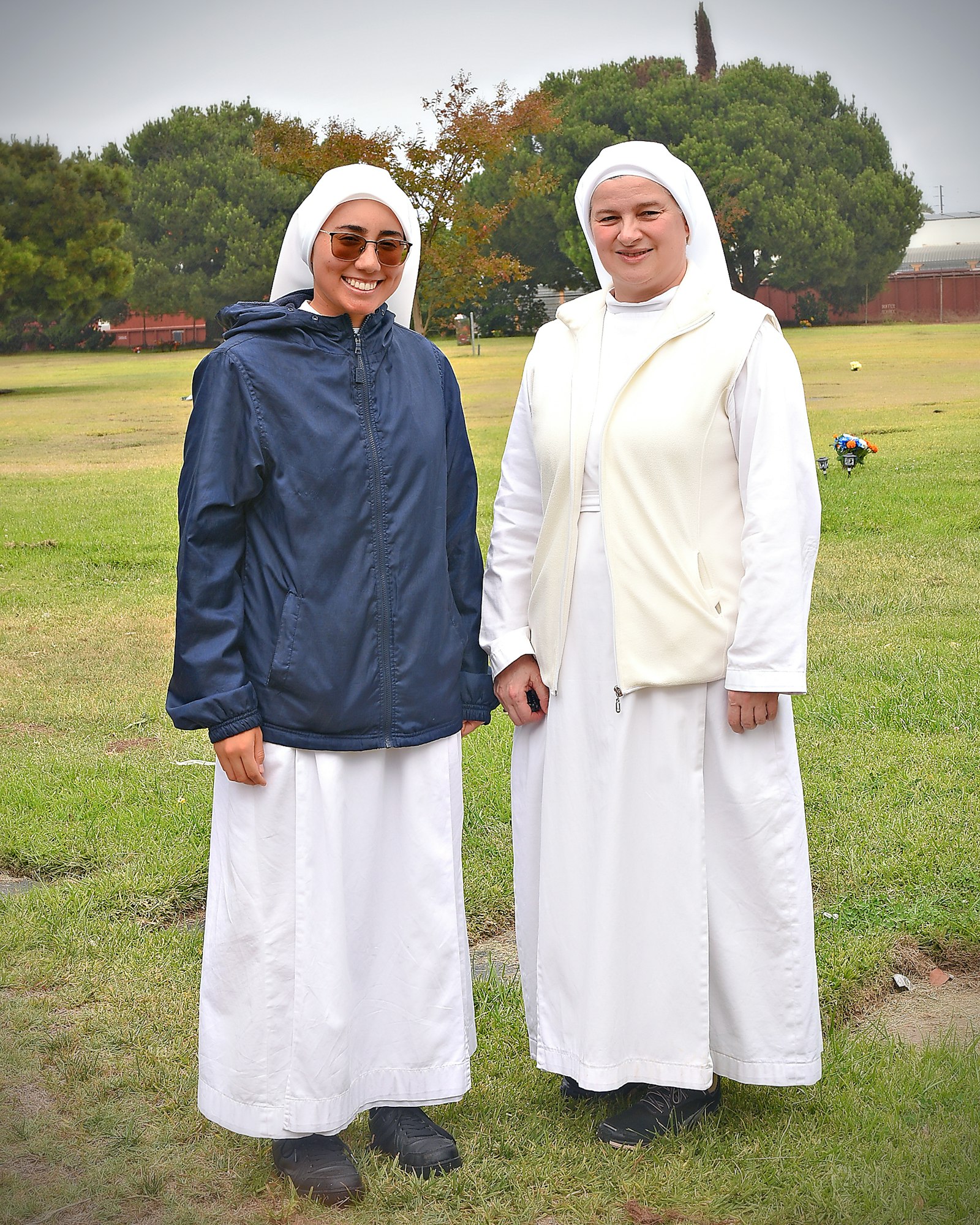 Two people wearing religious habits, possibly nuns, standing outside on grass.