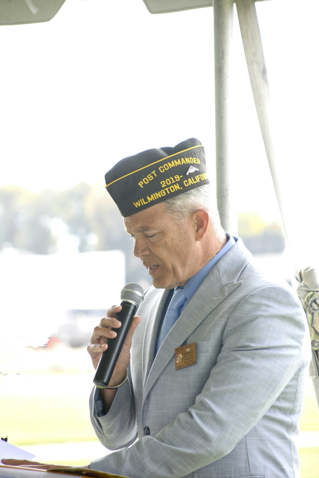 Man in a suit and service cap speaking into a microphone, possibly at a formal or military-related event.
