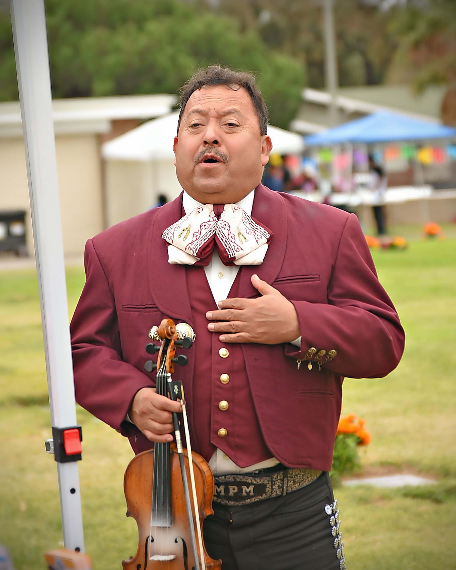 A musician in traditional attire holding a violin, possibly singing or talking. Outdoor setting.