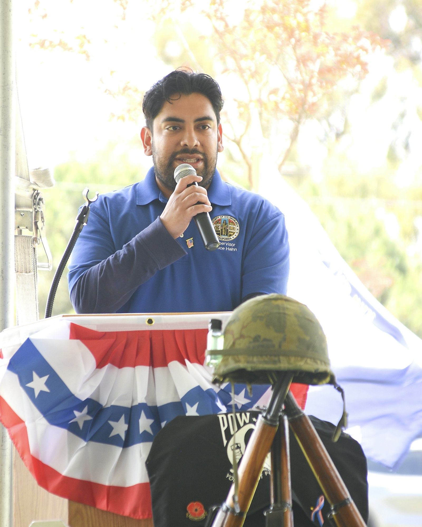 A man speaks into a microphone at a podium adorned with patriotic decor, including a helmet and rifles as a memorial display.