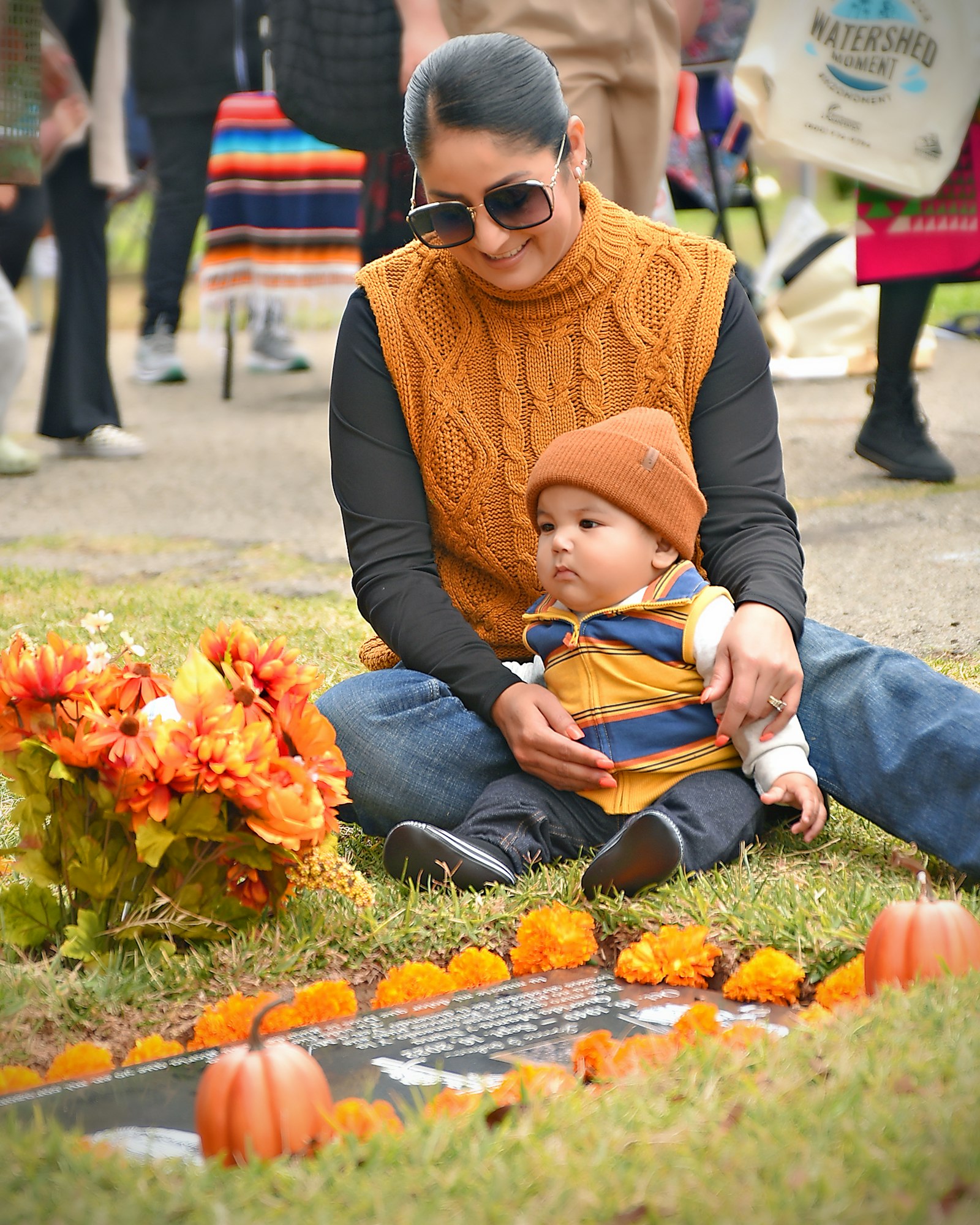 A woman in sunglasses and a sweater with a baby on her lap, sitting by a gravestone adorned with flowers and marigolds.