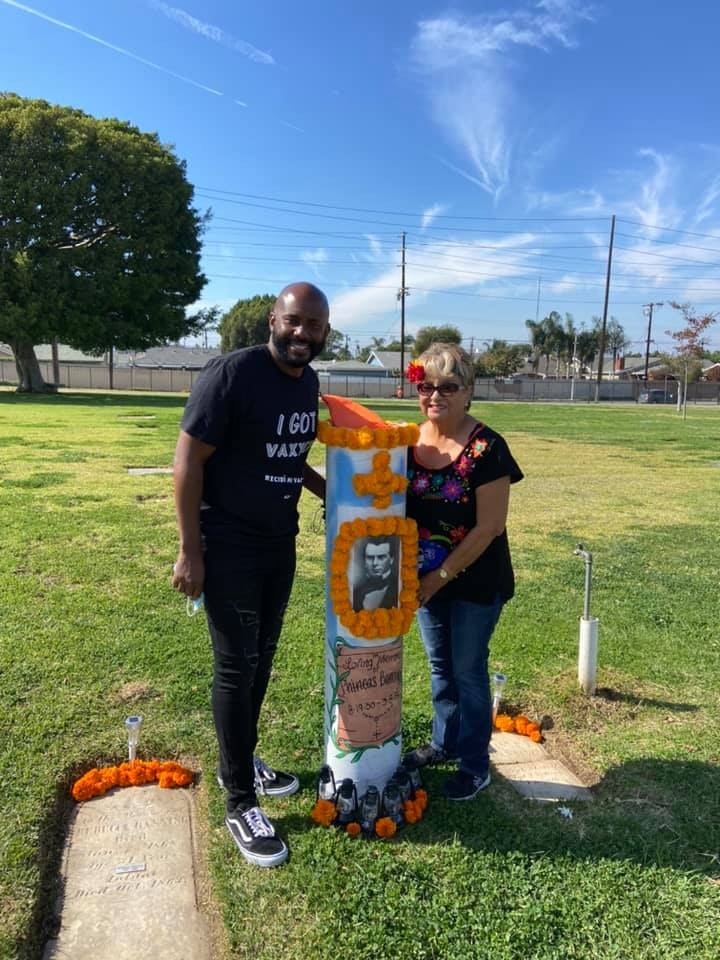 A man and woman pose by a decorated grave marker in a cemetery, commemorating a loved one with flowers and a photo.