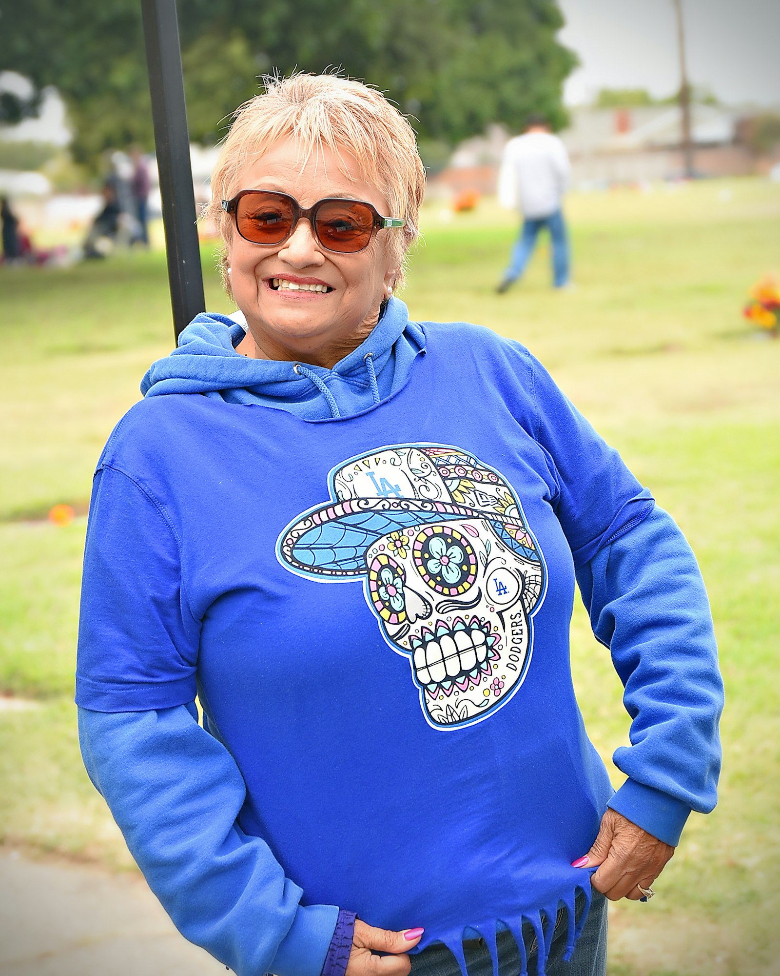 A smiling person in sunglasses wearing a blue hoodie with a colorful skull design, standing outdoors.