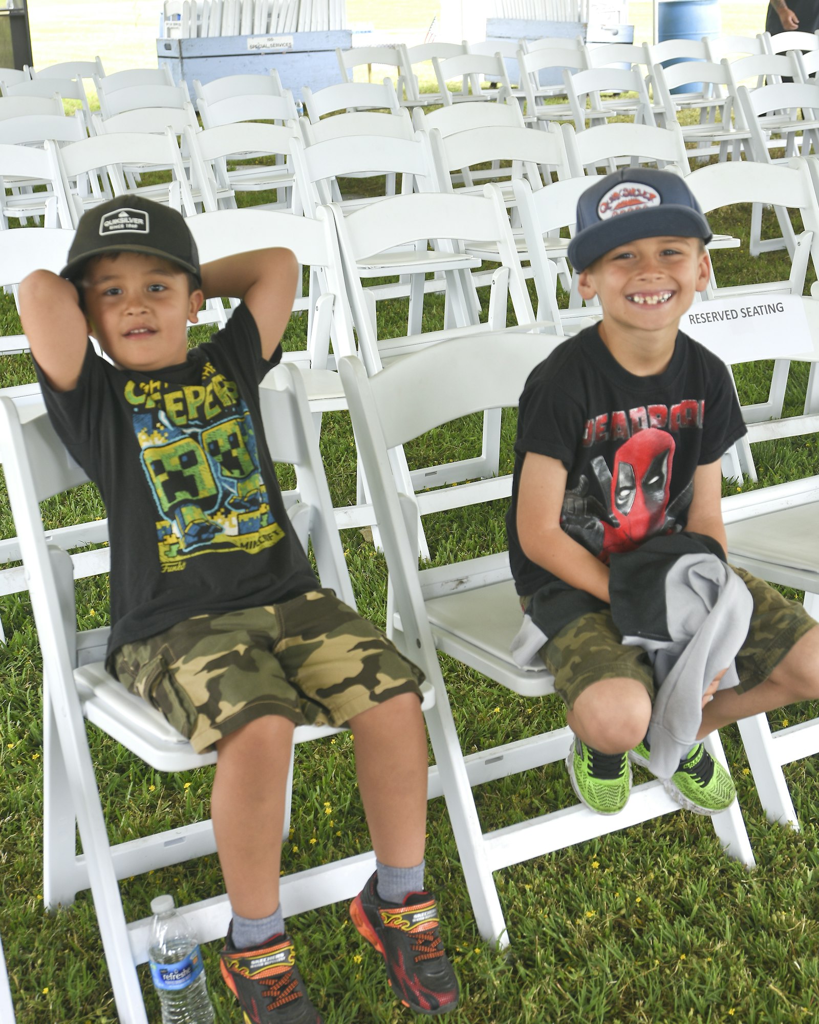 May contain: baseball cap, cap, clothing, hat, t-shirt, shorts, grass, plant, face, head, person, photography, portrait, footwear, shoe, chair, furniture, boy, child, male, lawn, people, and sitting