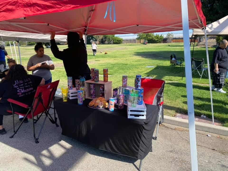 A market scene under a red tent with people, colorful drinks on display, and snacks like cookies on a table in a park setting.