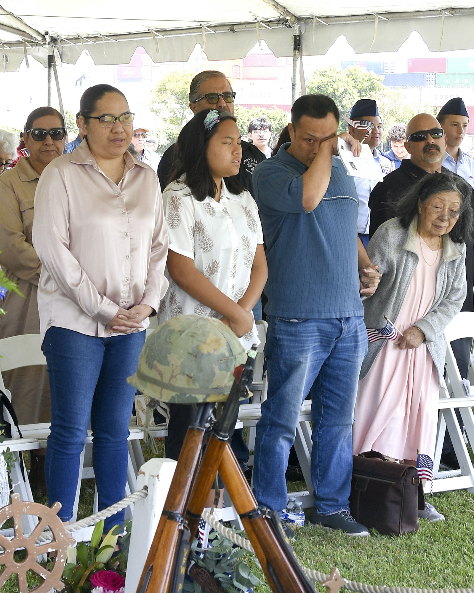 A group of solemn people at an event with a ceremonial rifle and helmet display, possibly a military memorial service.