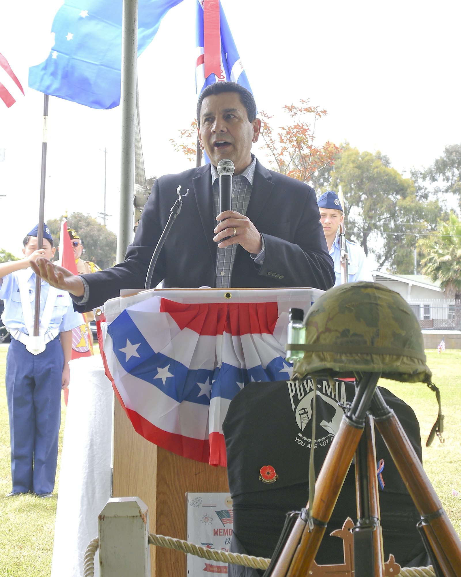 Man speaking at podium with microphone, flags, military helmet, and POW-MIA symbol in a commemorative setting.