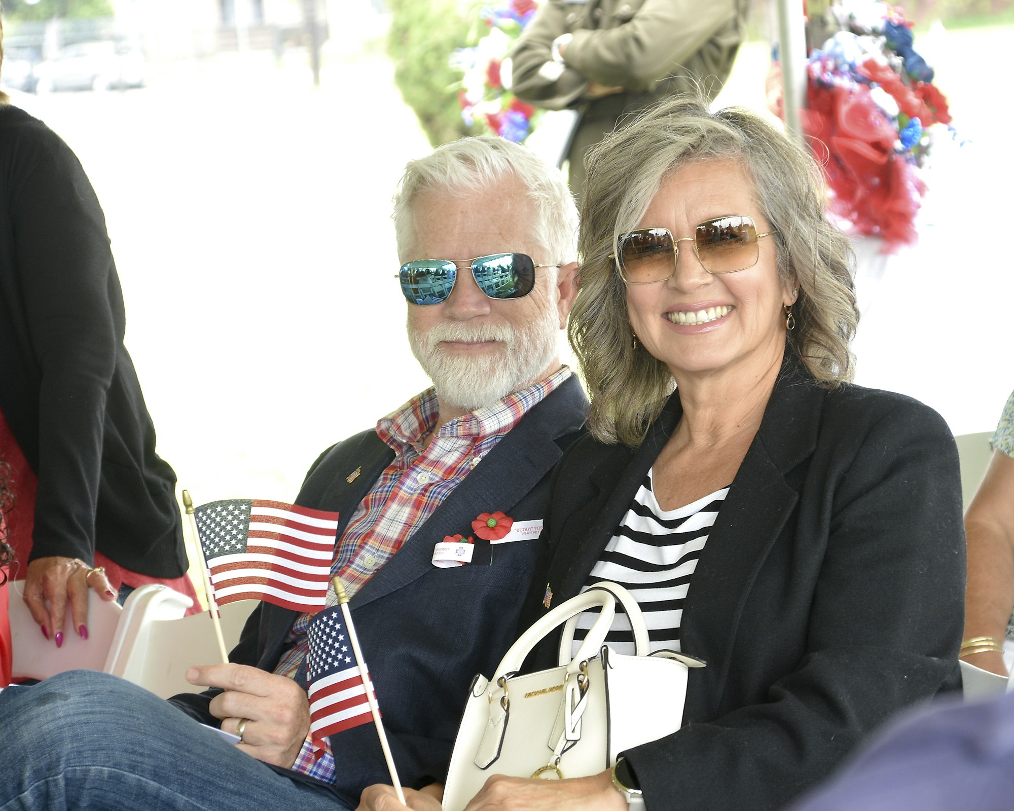 A smiling man and woman seated together, holding small American flags, adorned with remembrance poppies, at an outdoor event.