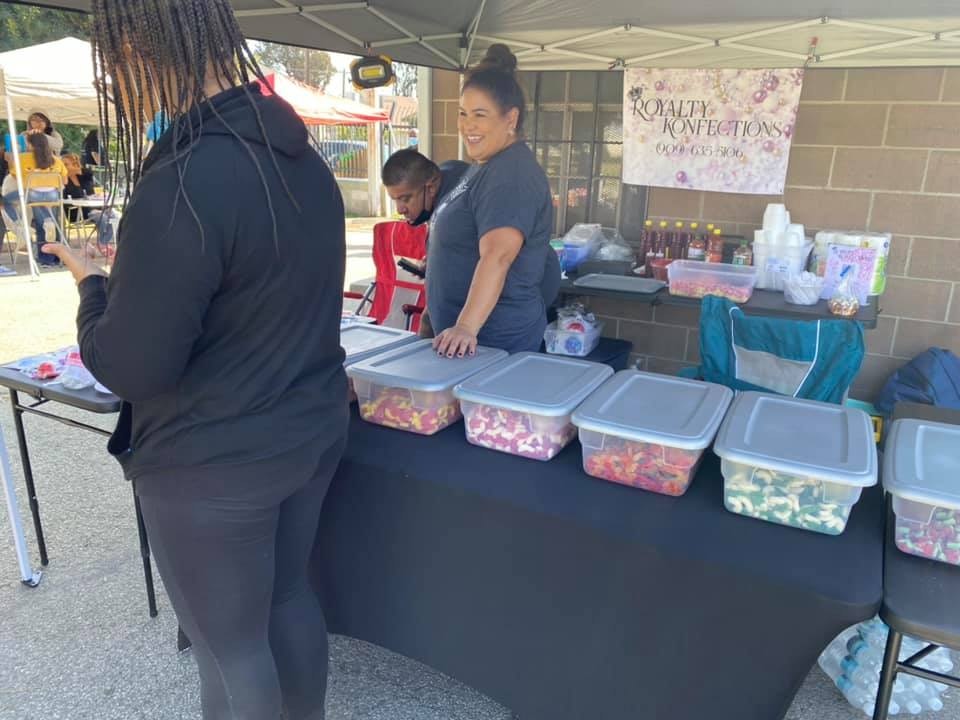 A vendor booth with a smiling woman, colorful containers of sweets, and customers at an outdoor event.