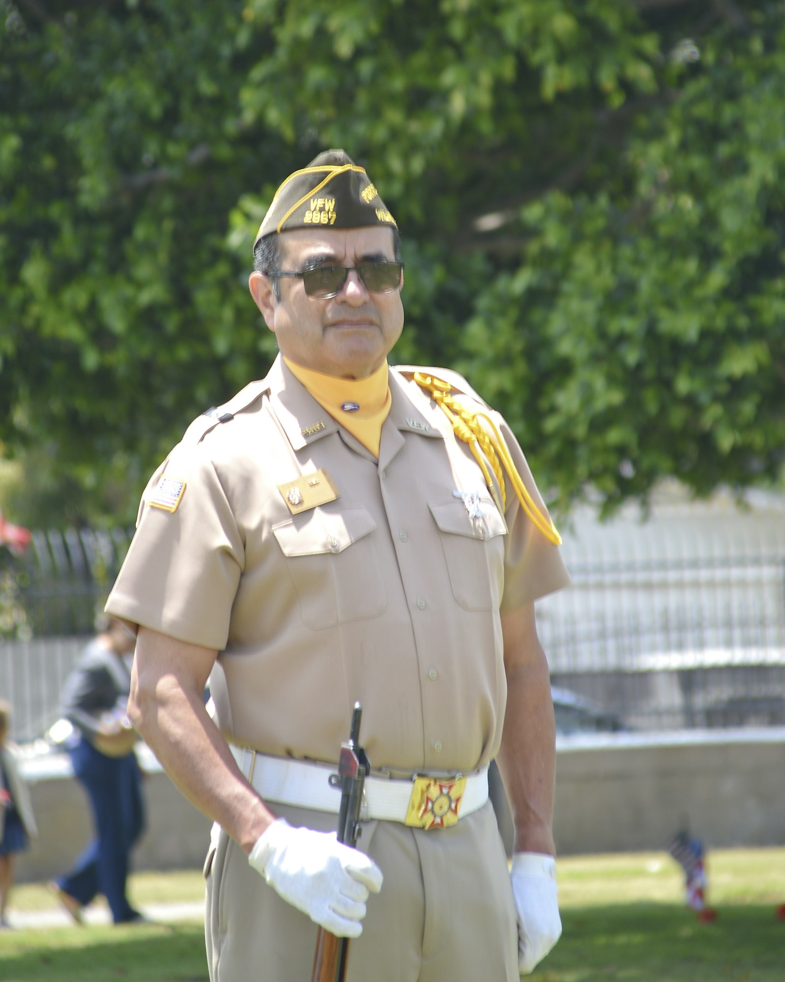 A person in a military-style uniform with a cap, holding a rifle, and wearing white gloves, standing outdoors.