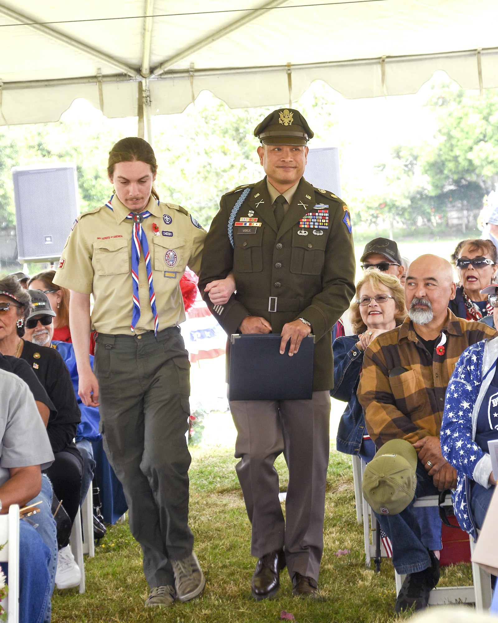 A Boy Scout and a military officer walking side by side at an event with onlookers in the background.