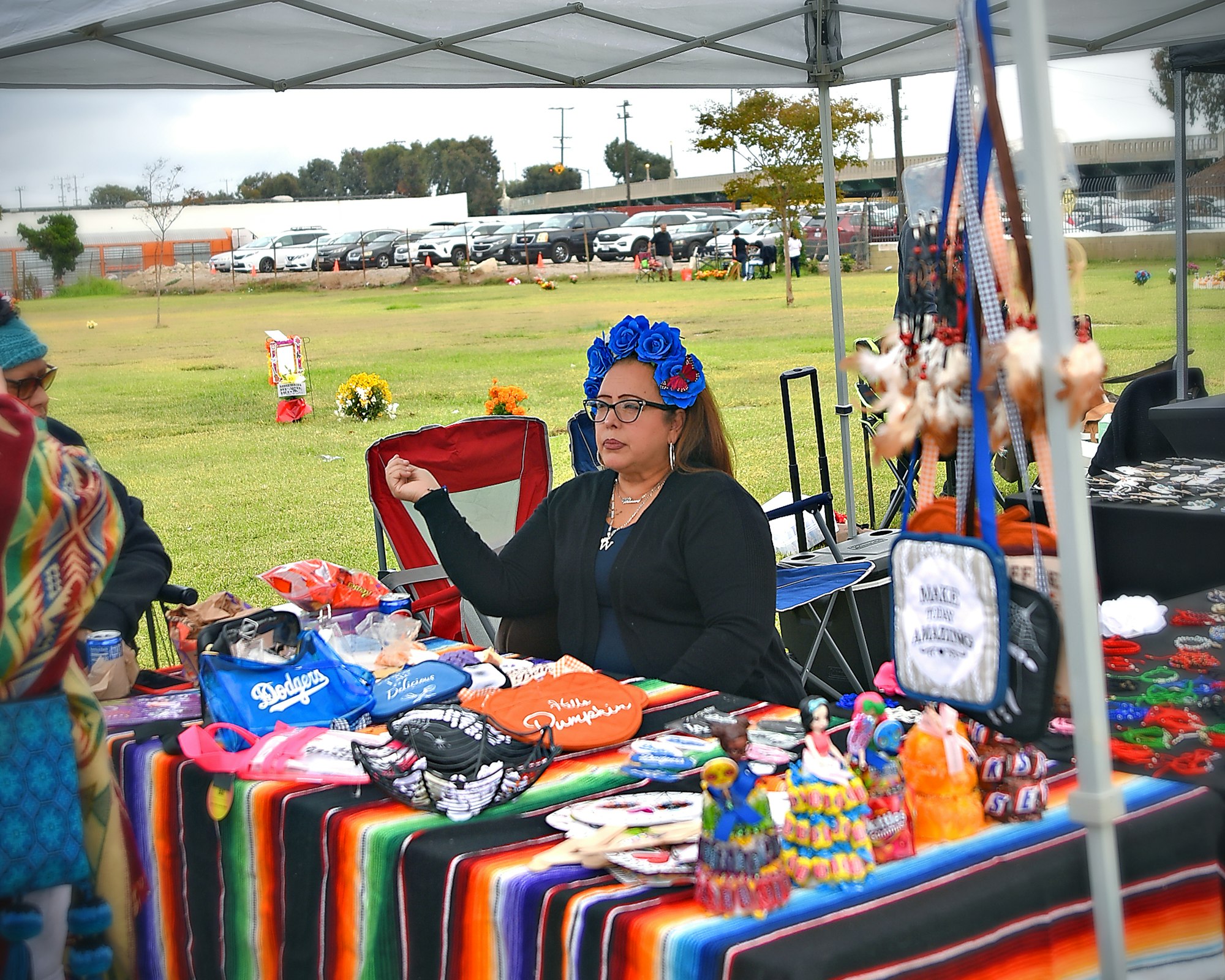 A woman with blue floral headwear sits at an outdoor market stall featuring colorful textiles and assorted goods.