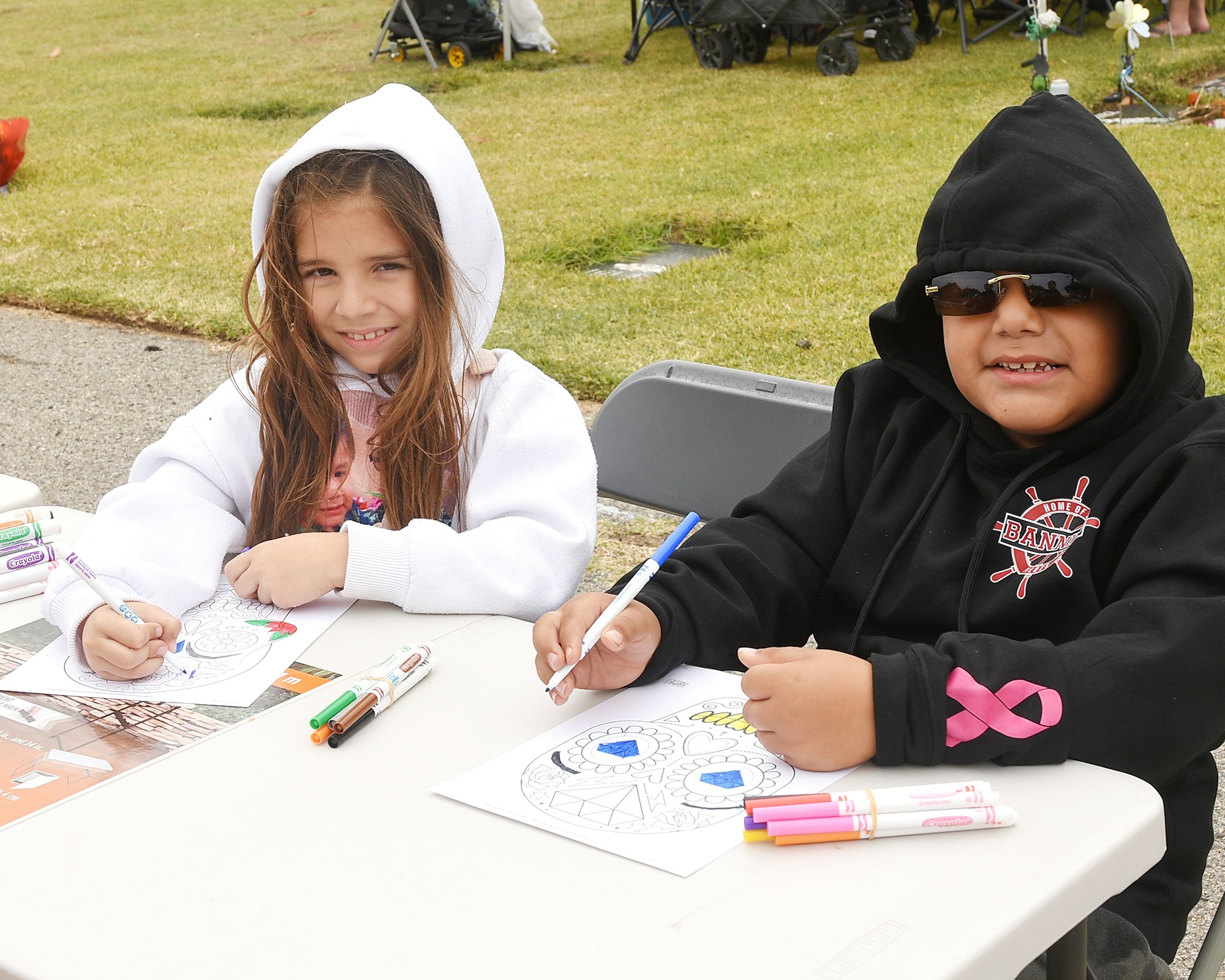 Two kids coloring at a table outdoors, smiling, with markers and coloring sheets.