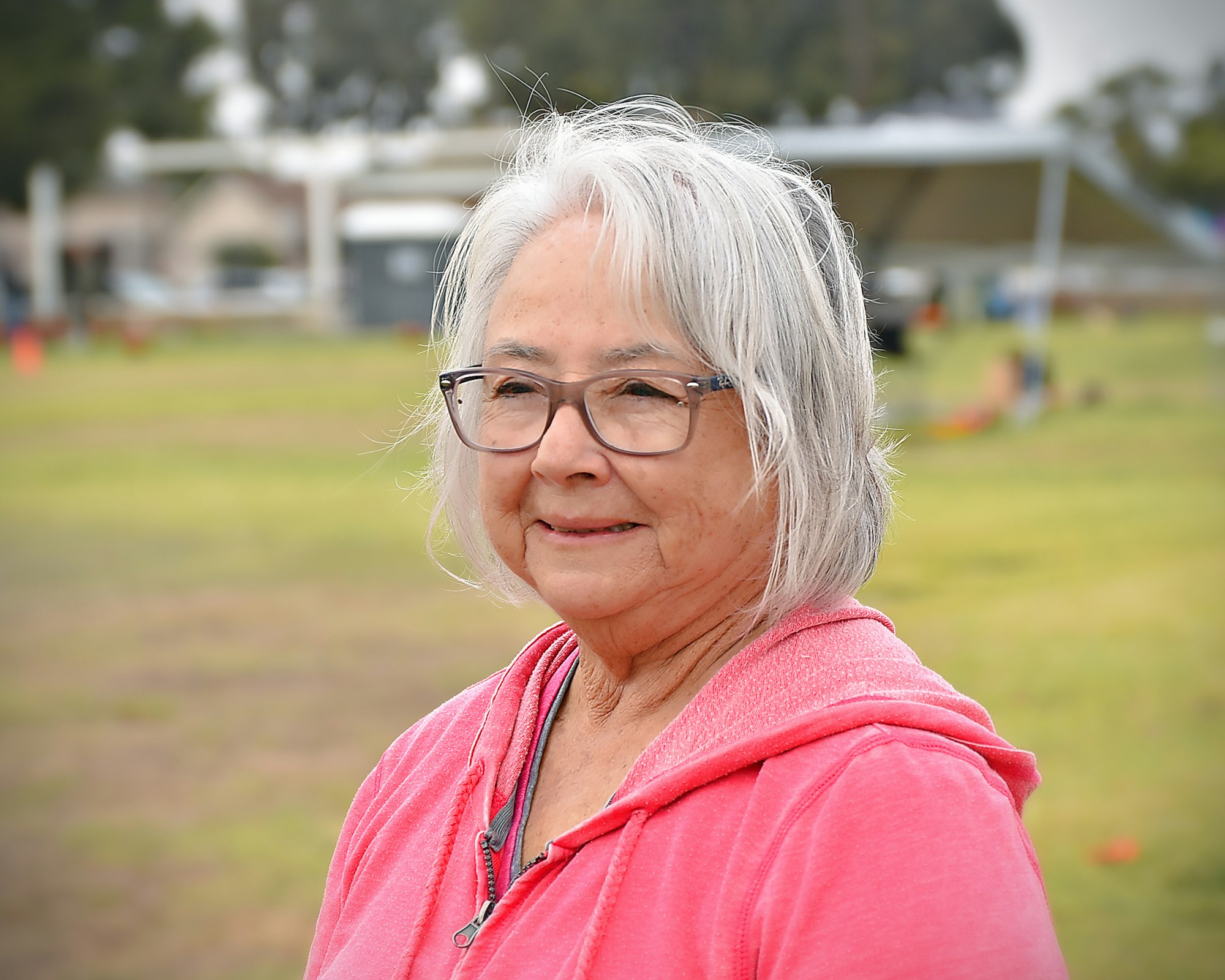 An elder woman with glasses and a pink hoodie, slightly smiling outdoors.