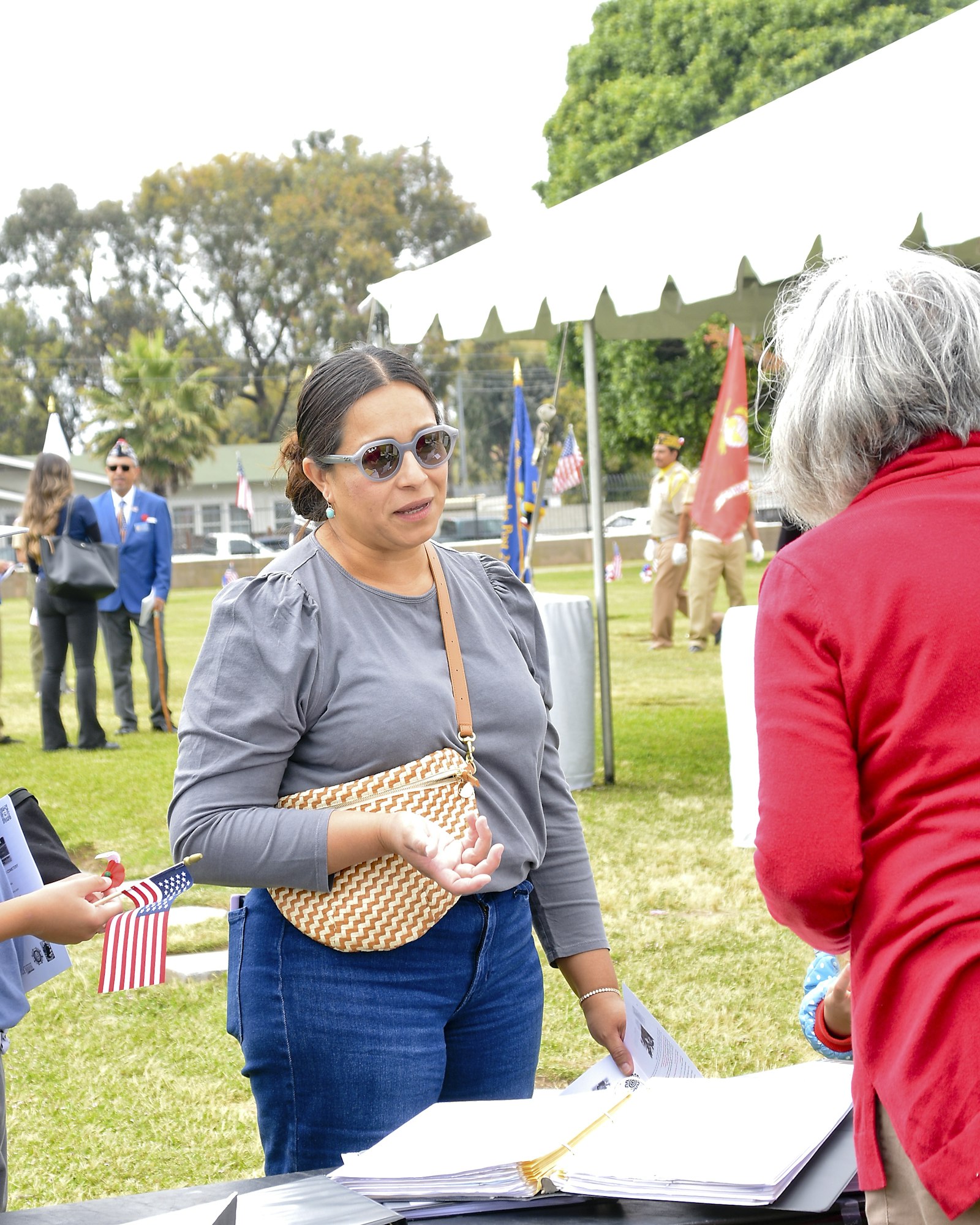 A woman conversing at an outdoor event with tents and flags, a child holding an American flag nearby.