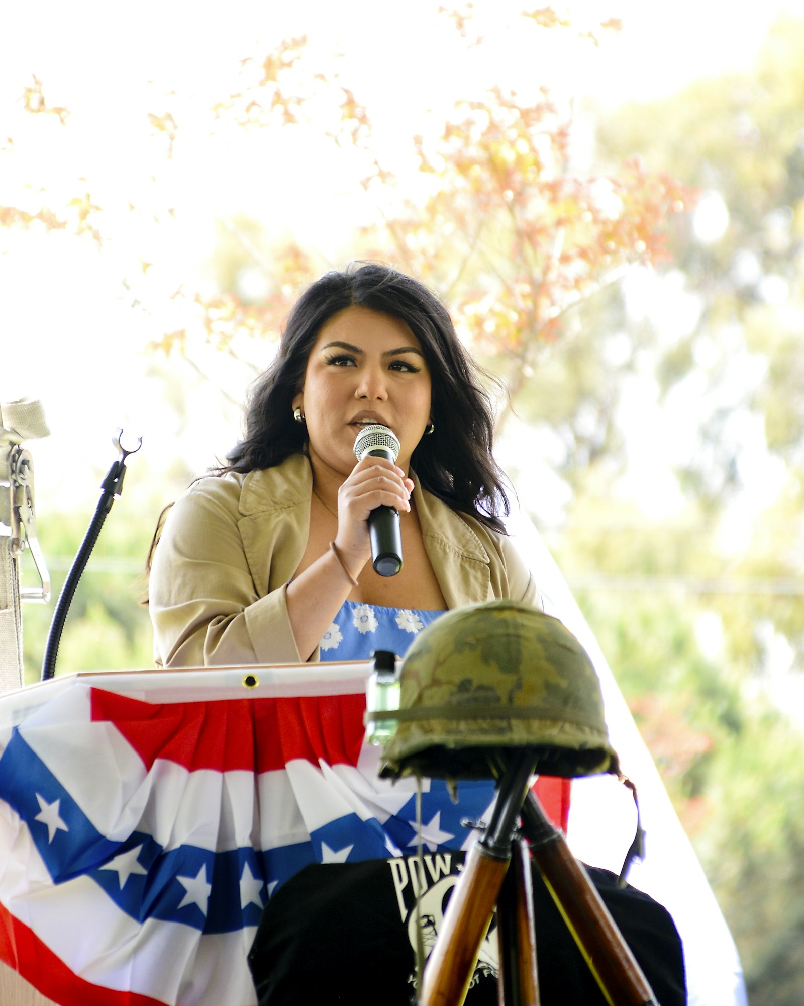 A person speaking into a microphone at a podium with an American flag, a POW/MIA flag, and a military helmet.