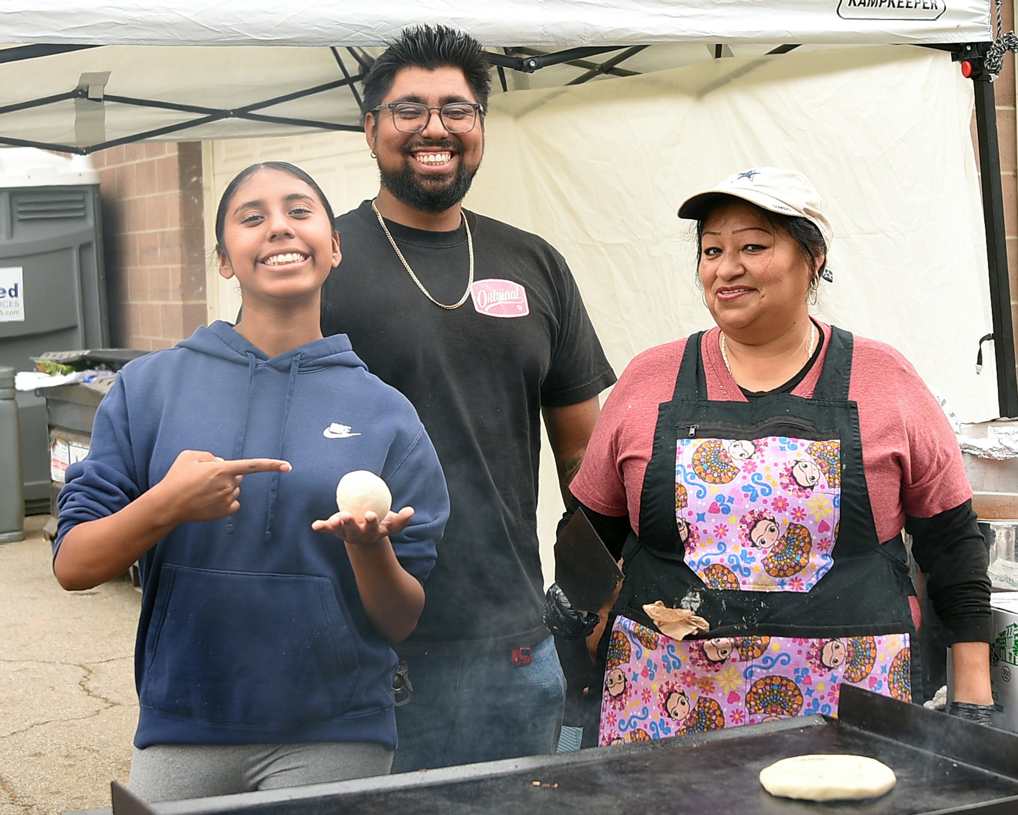 Three people at an outdoor event, one holding a tortilla, with a cooking area and warm smiles.