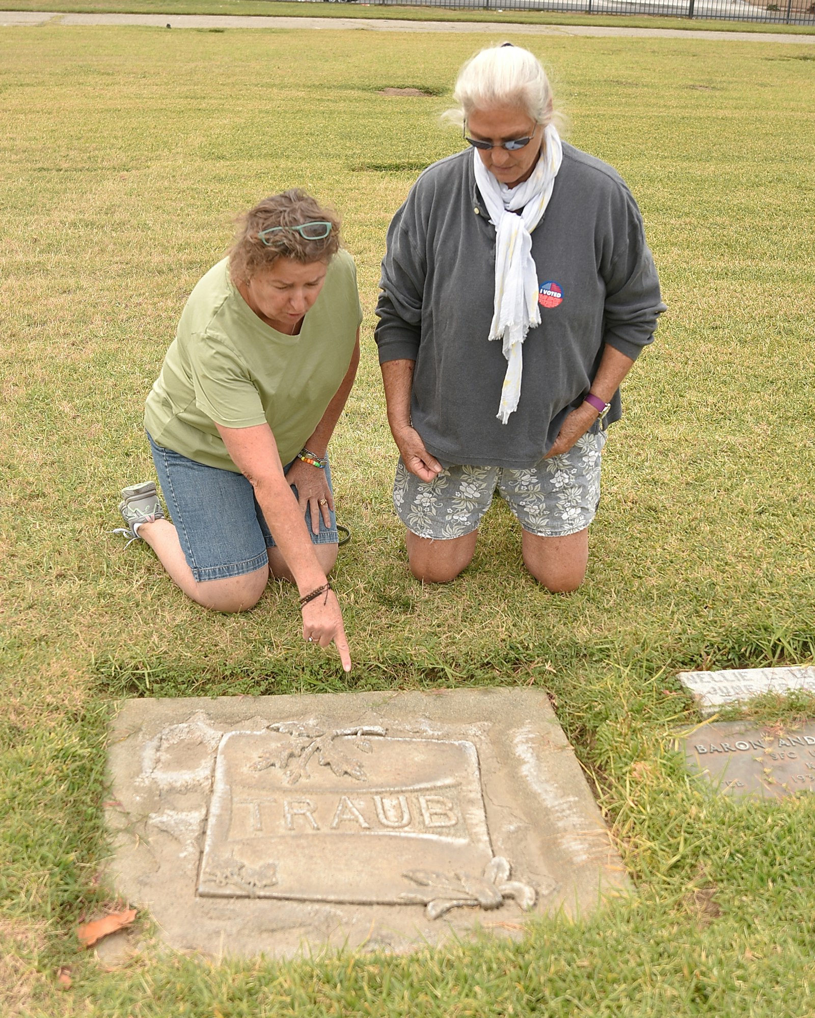 Two people examining a gravestone marked "TRAUB" in a grassy area.