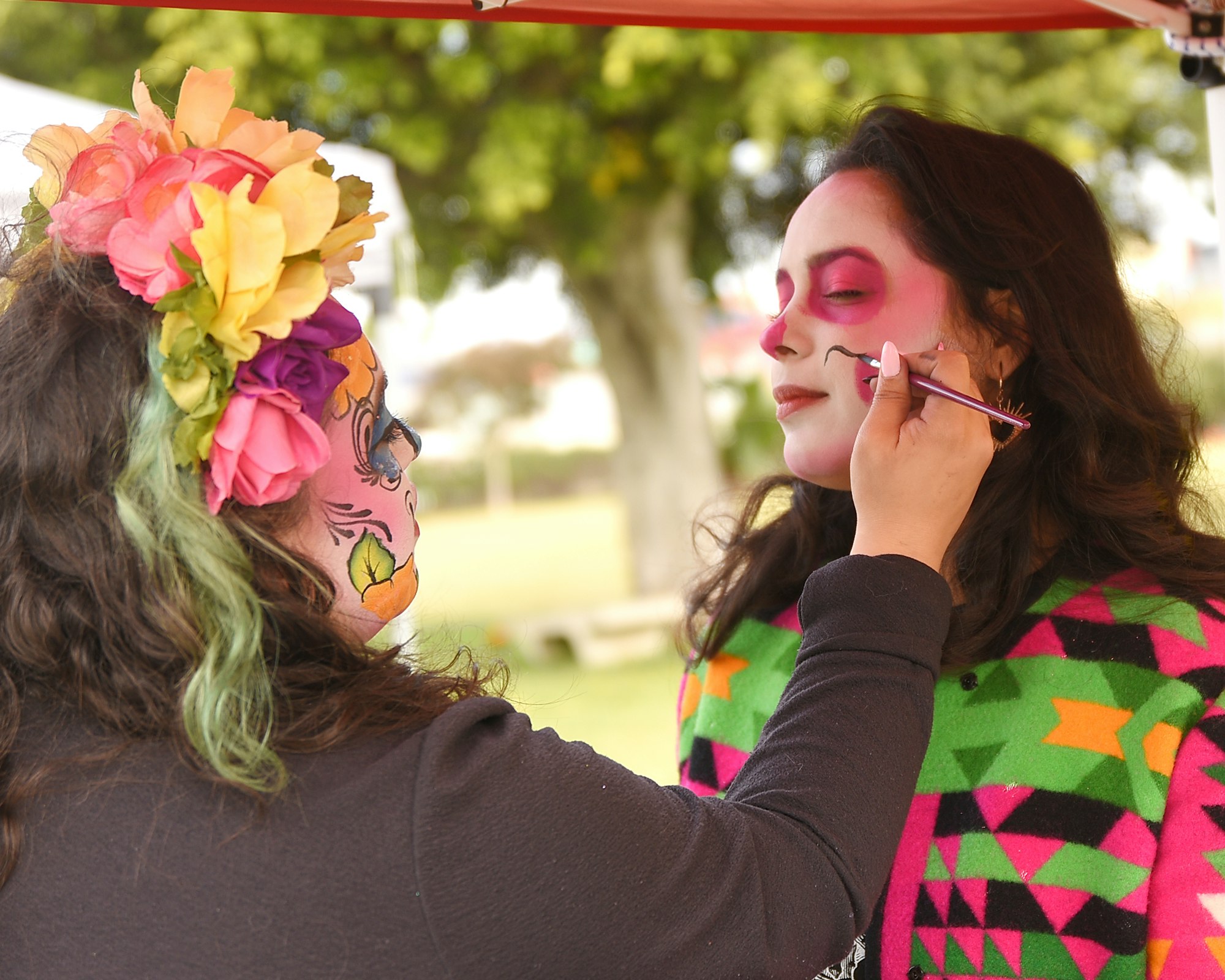 Face painting session in action, one person with a flower crown and colorful makeup being applied by another.