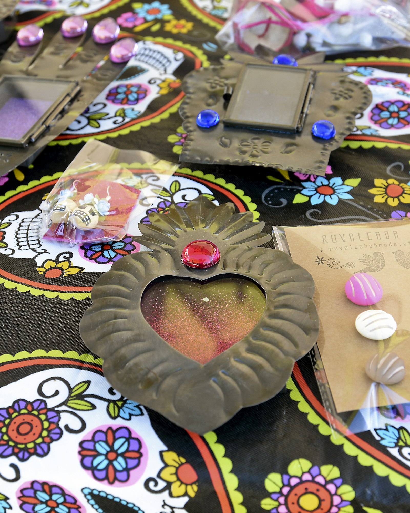 Close-up of a Day of the Dead-themed craft display. The table features small decorative metal frames, including a heart-shaped frame with a red jewel at the top and several rectangular frames with colorful accents. The background is a vibrant tablecloth patterned with colorful sugar skulls and floral motifs. Small craft supplies, such as buttons and embellishments, are also visible in clear plastic bags.