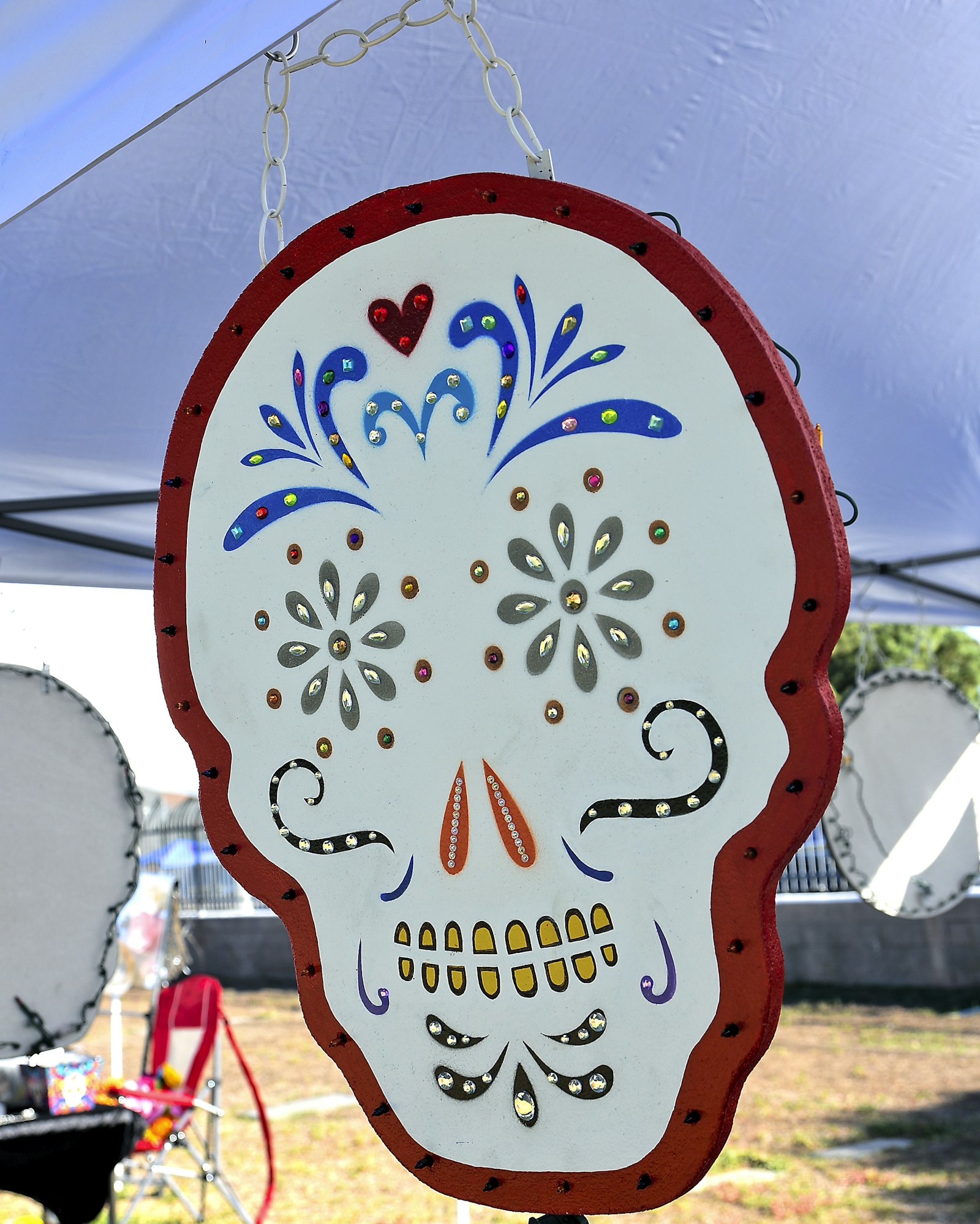 Decorative Day of the Dead sugar skull art piece hanging from a chain under a canopy. The skull is white with a red border and features colorful painted details in blue, gold, and red, including floral designs, hearts, and stylized facial features. In the background, there is a red chair, part of another similar skull decoration, and an outdoor event setting.