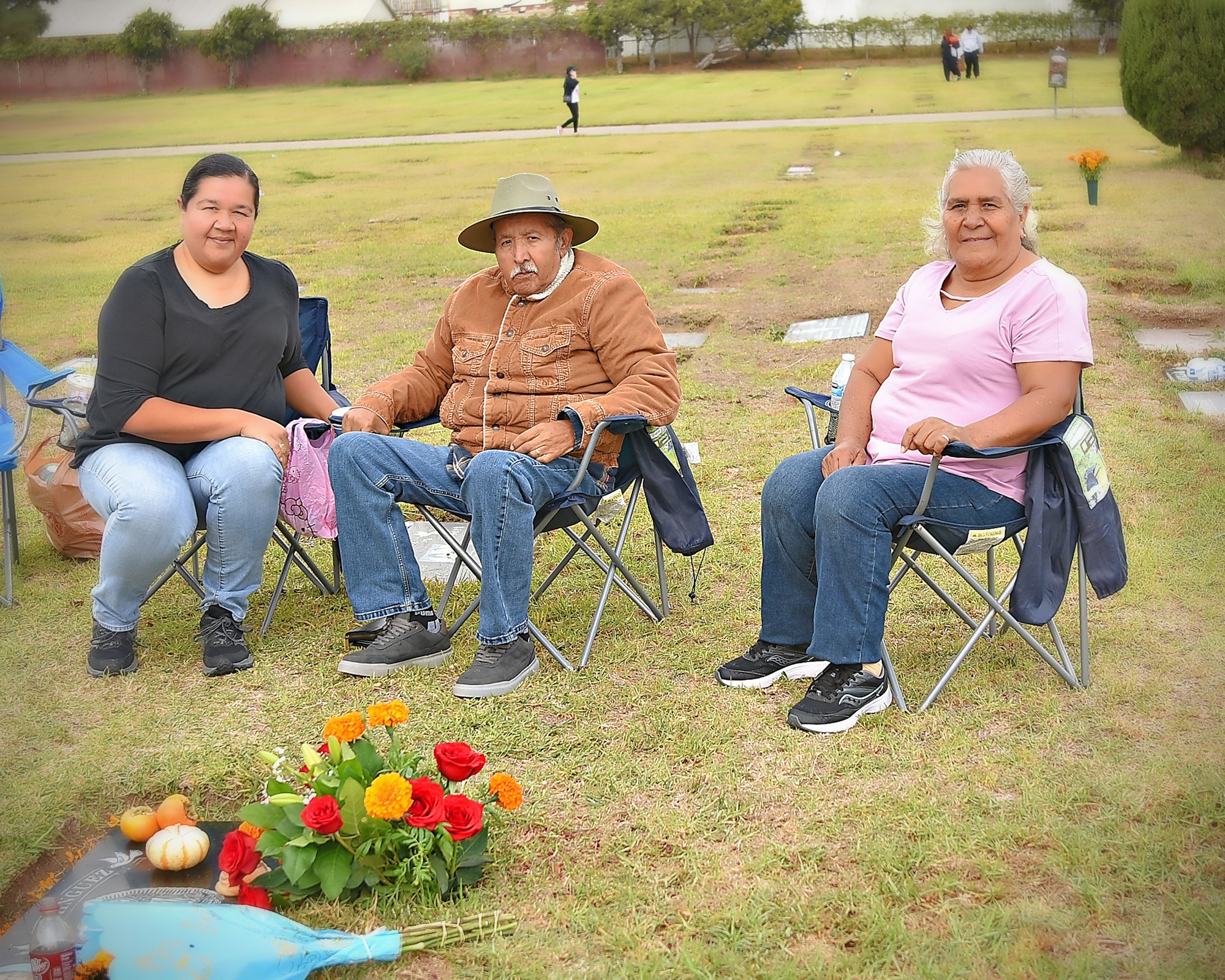 Three people seated on folding chairs at a gravesite with flowers, on a cloudy day.
