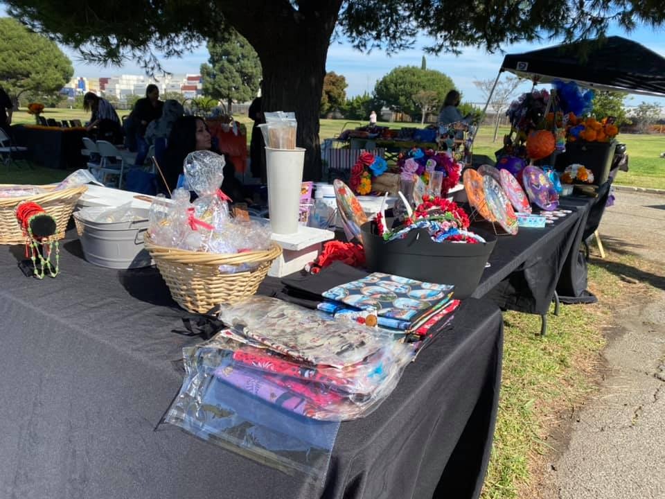 A table at an outdoor market displaying various crafts, decorations, and colorful items, with people in the background.