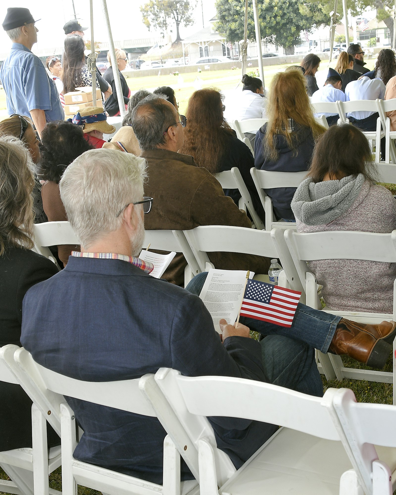 People sitting on chairs at an outdoor event with one person holding a pamphlet and an American flag.