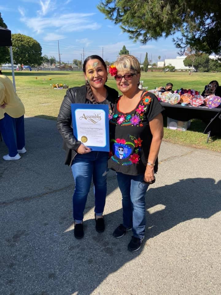 Two women pose together outdoors, one holding a certificate. The setting appears festive, with decorations visible in the background.