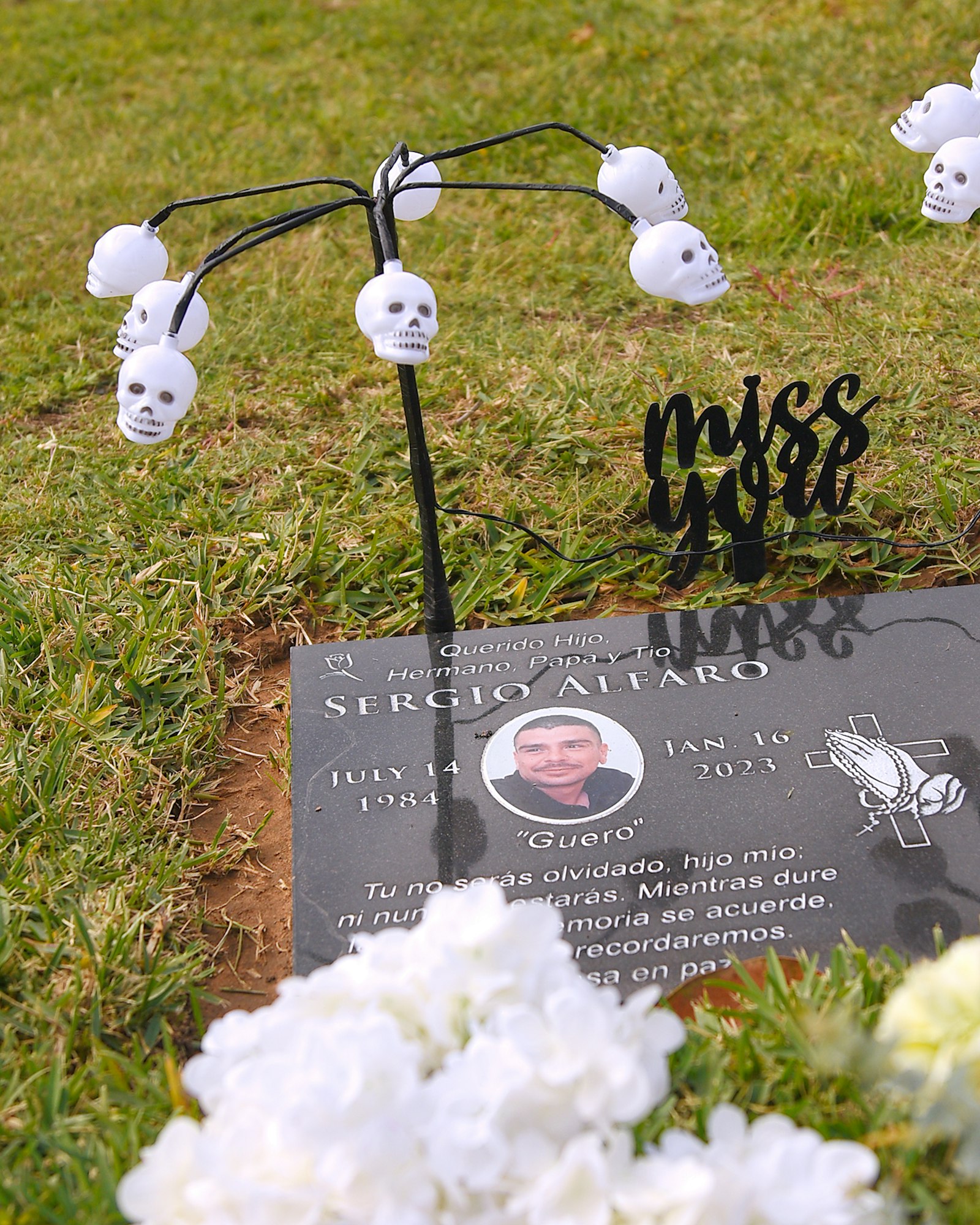 A grave marker with a photo, flowers, skull decorations, and a "miss you" sign.