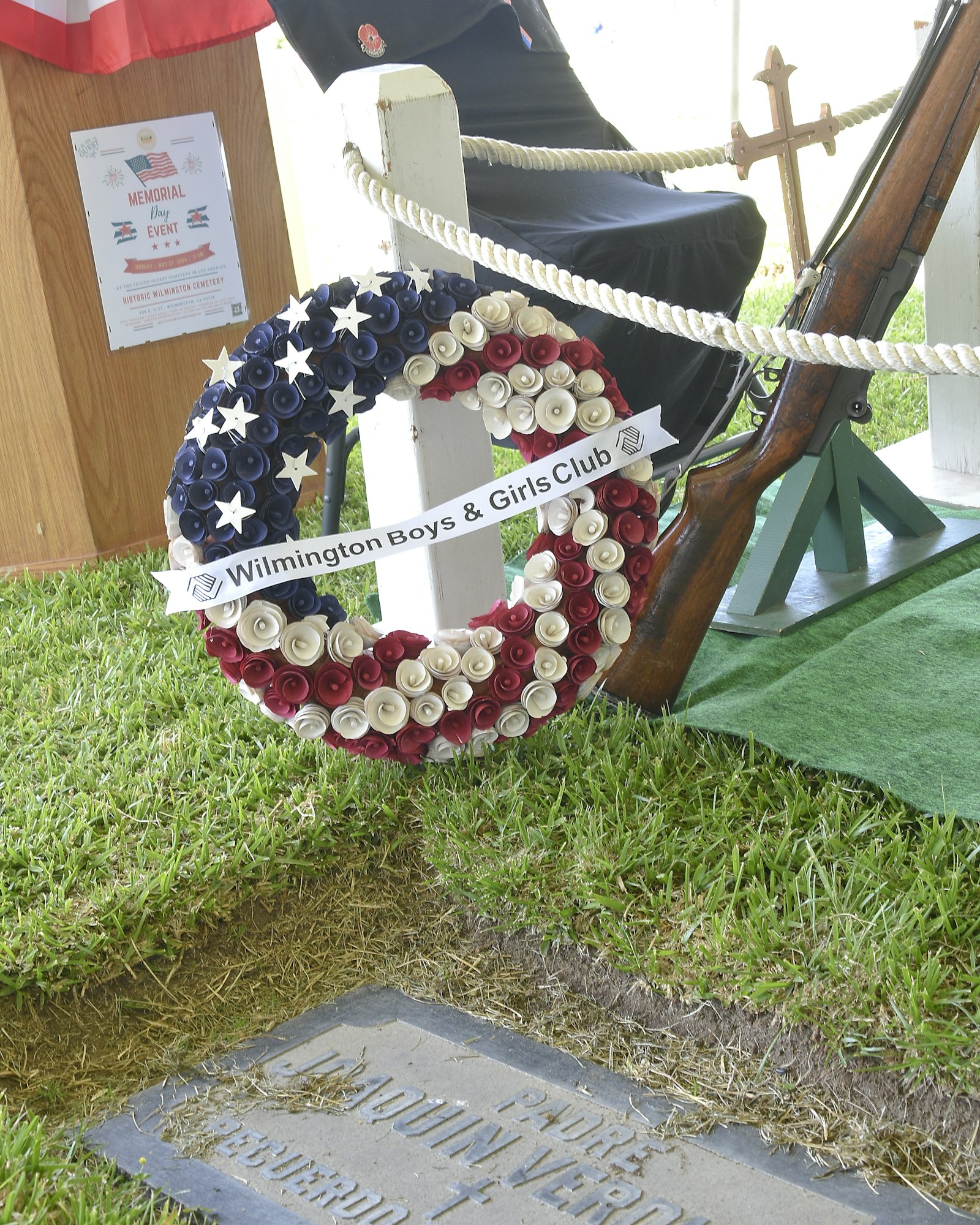 A wreath with red, white, and blue ribbons, a memorial event flyer, a folded chair, and a military rifle with bayonet, honoring veterans.