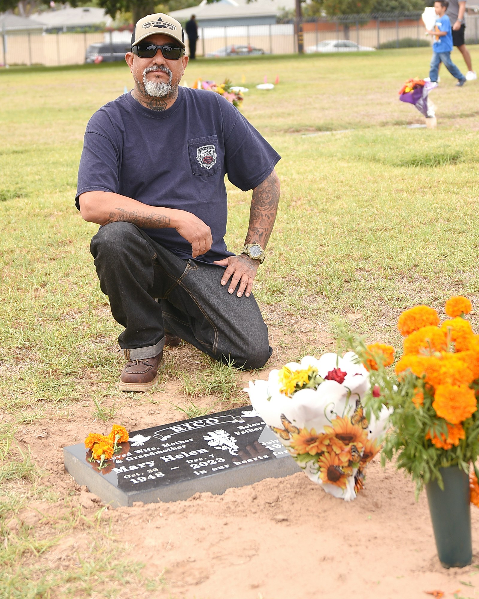 A man kneeling by a grave with flowers, outdoors on a grassy field, with children playing in the background.