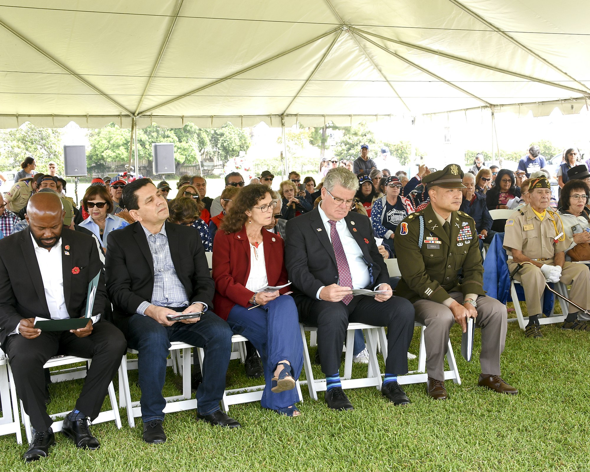 Group of people sitting in rows under a tent, likely at an outdoor event or ceremony.