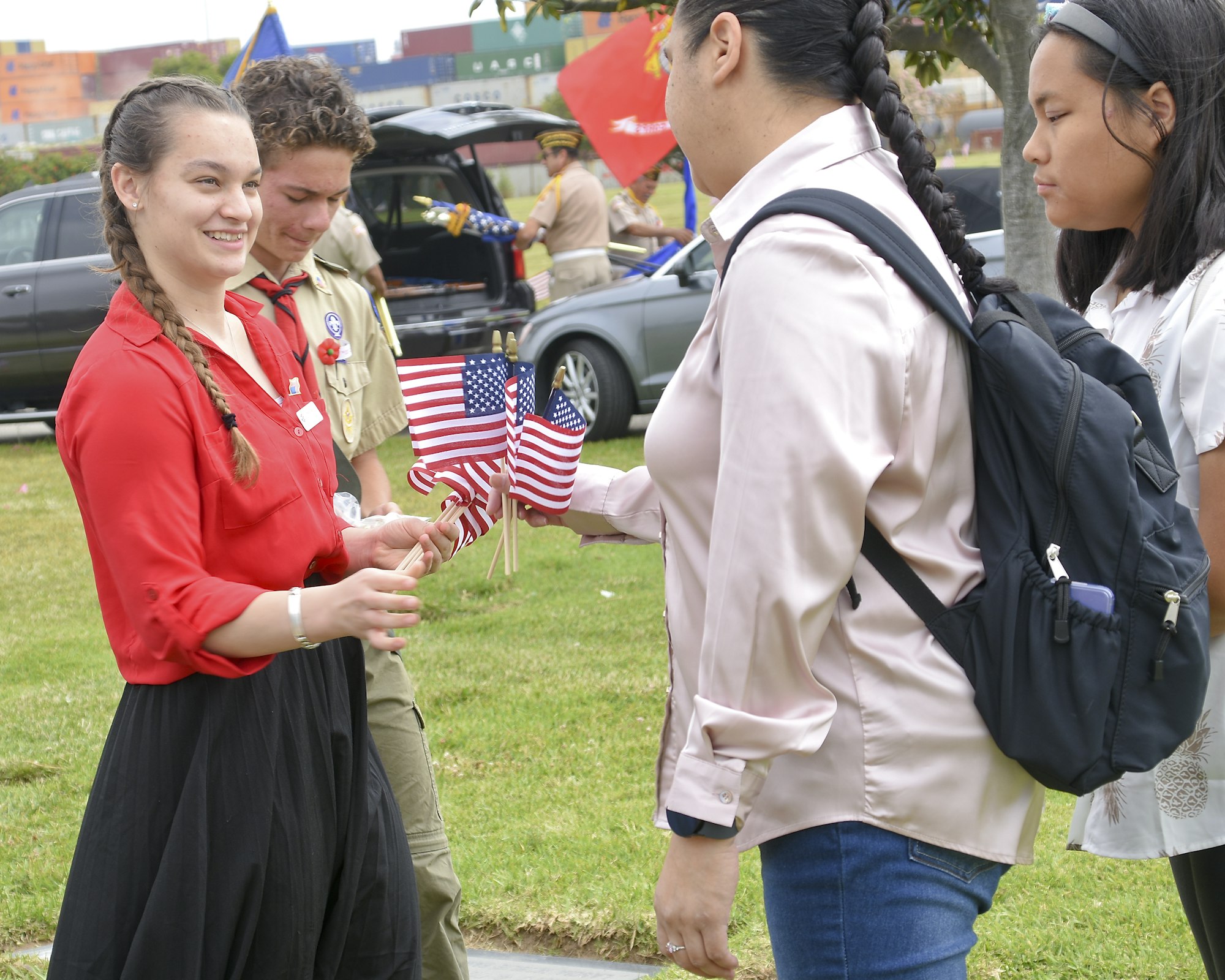 People exchanging small American flags; two in scout uniforms, one with a backpack, casual outdoor setting.